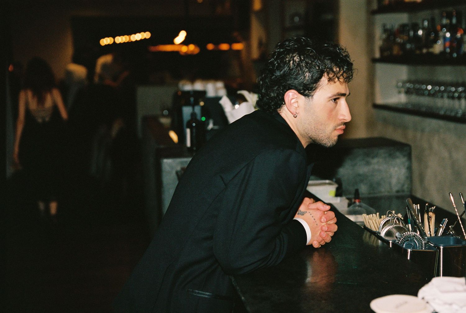 A man in a black suit leans on a bar counter at an evening event.