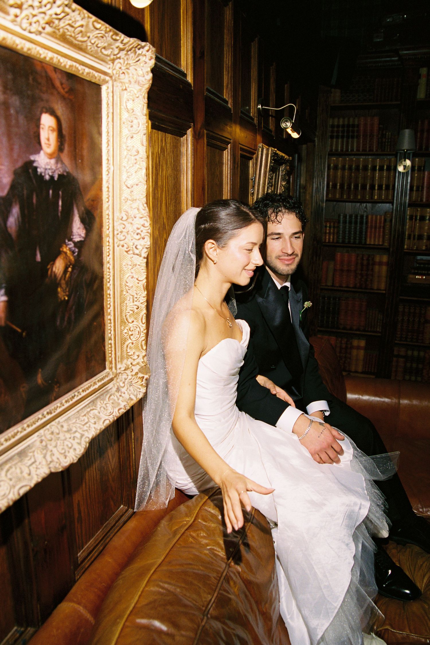 Bride and groom sitting on leather sofa in wood-paneled library with antique portrait painting.