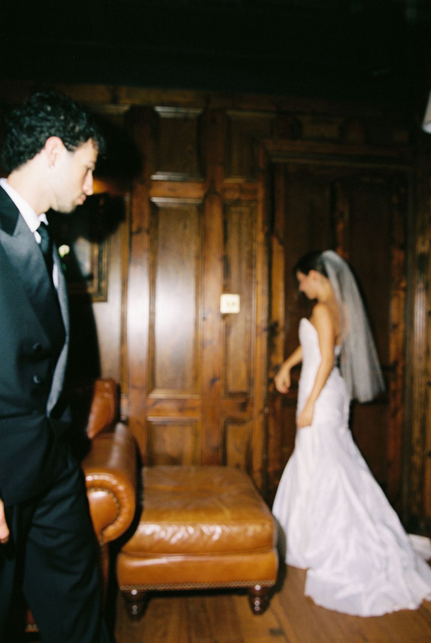 Bride in mermaid wedding dress and groom in tuxedo sharing first look in wood-paneled room.