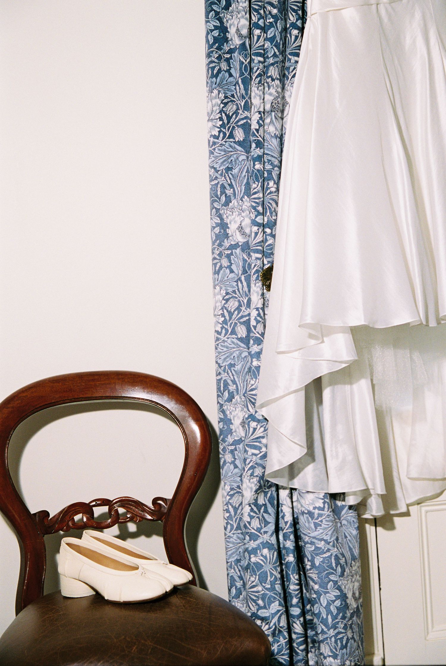 White wedding dress hanging beside blue floral curtains with white block-heel bridal shoes on a Victorian chair.