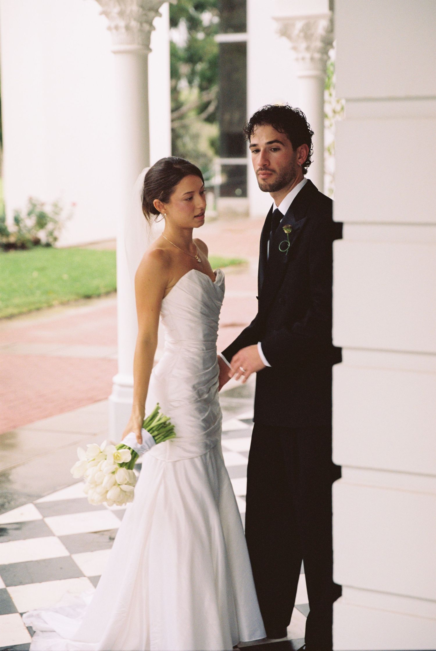 Bride in strapless mermaid gown holds white bouquet beside groom in black suit at columned venue.