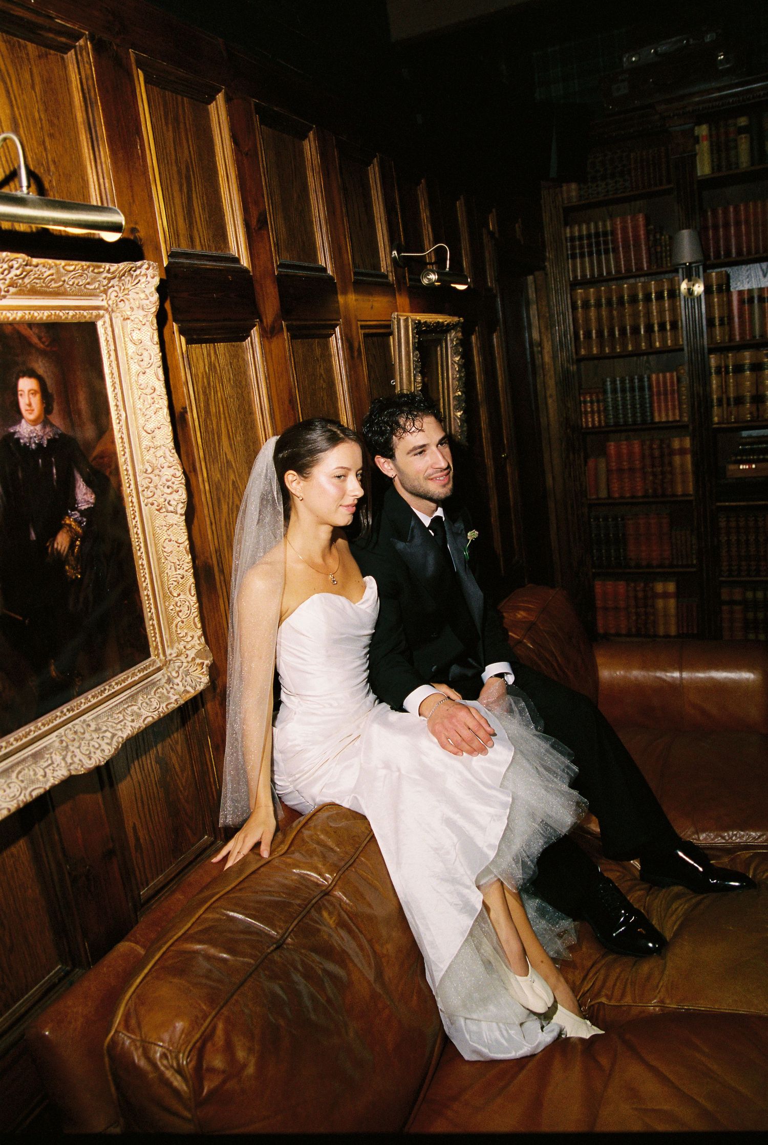 Bride and groom sit together on leather sofa in wood-paneled library with antique portraits.