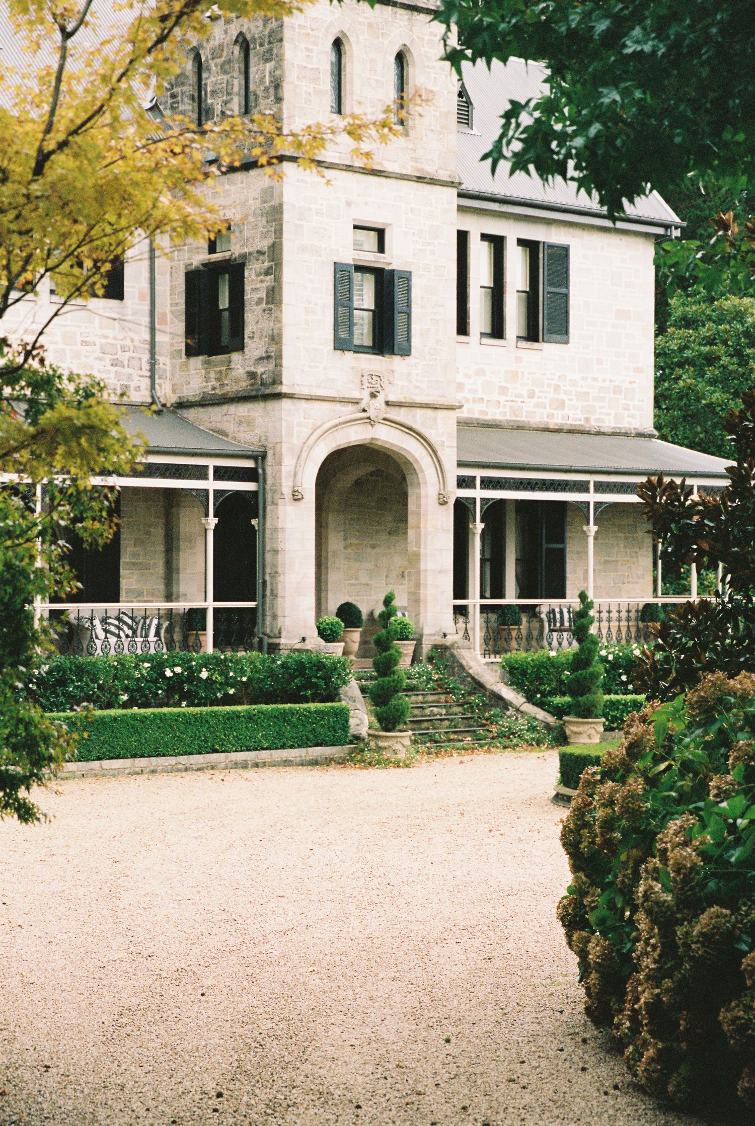 Historic stone manor house with arched entrance, manicured hedges, and gravel driveway surrounded by lush greenery.