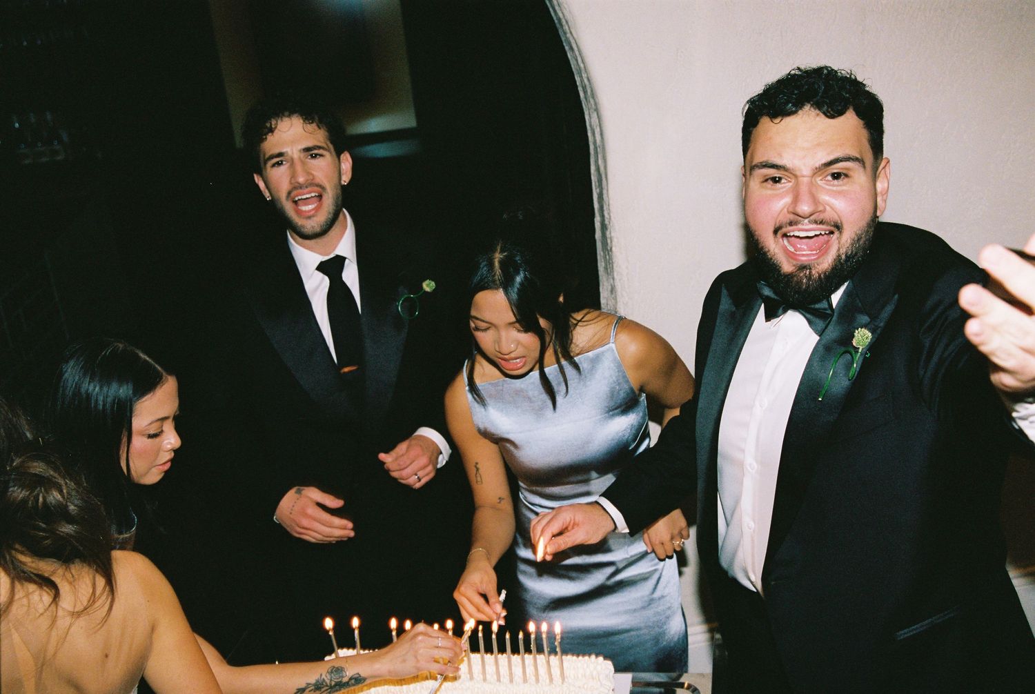 Wedding guests smiling and celebrating around a white cake with candles at a formal reception.