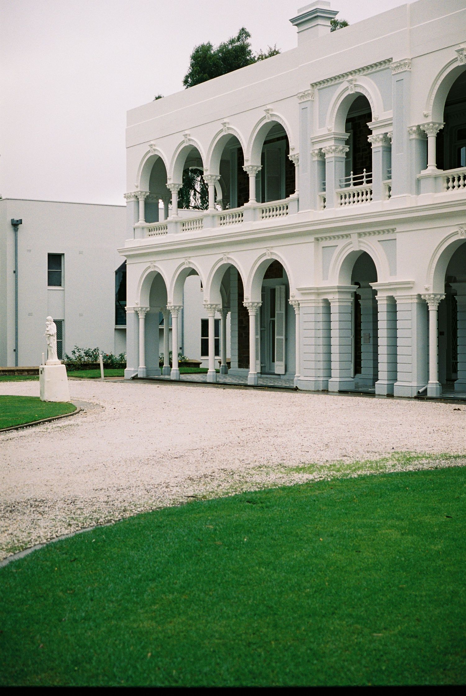 Elegant white colonial building with two-story arched colonnades, balustrades, and manicured green lawn on overcast day.