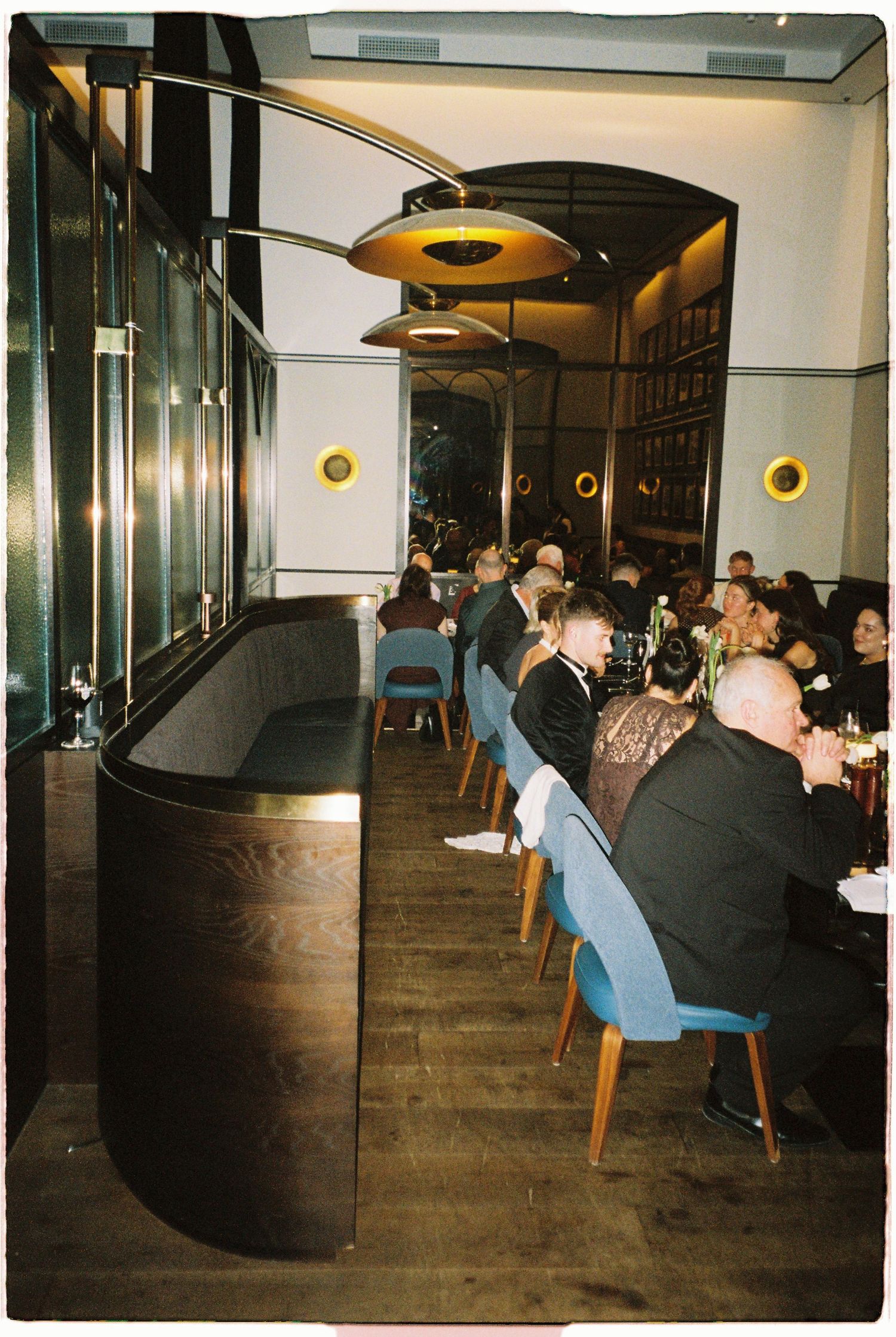 Wedding reception guests dining in a sophisticated restaurant with curved booths and warm pendant lighting.