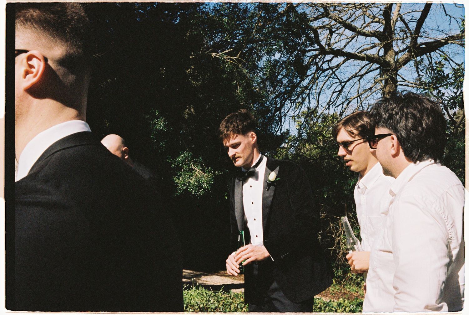 Groomsmen in black tuxedos and white shirts converse outdoors at a wedding ceremony surrounded by trees.