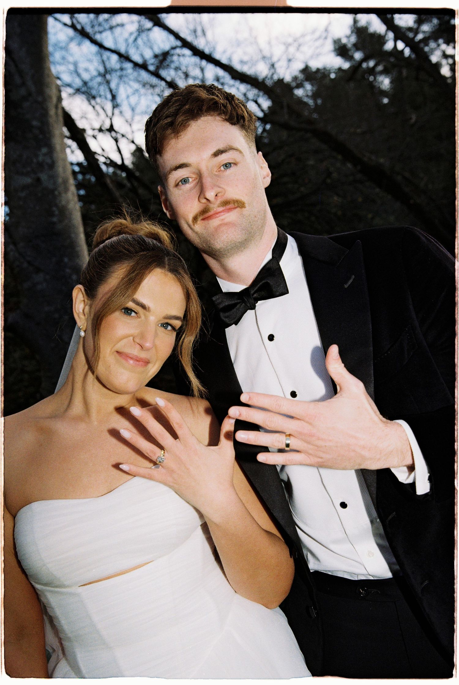 A newlywed couple proudly displays their wedding rings while posing together in a black tuxedo and white gown.