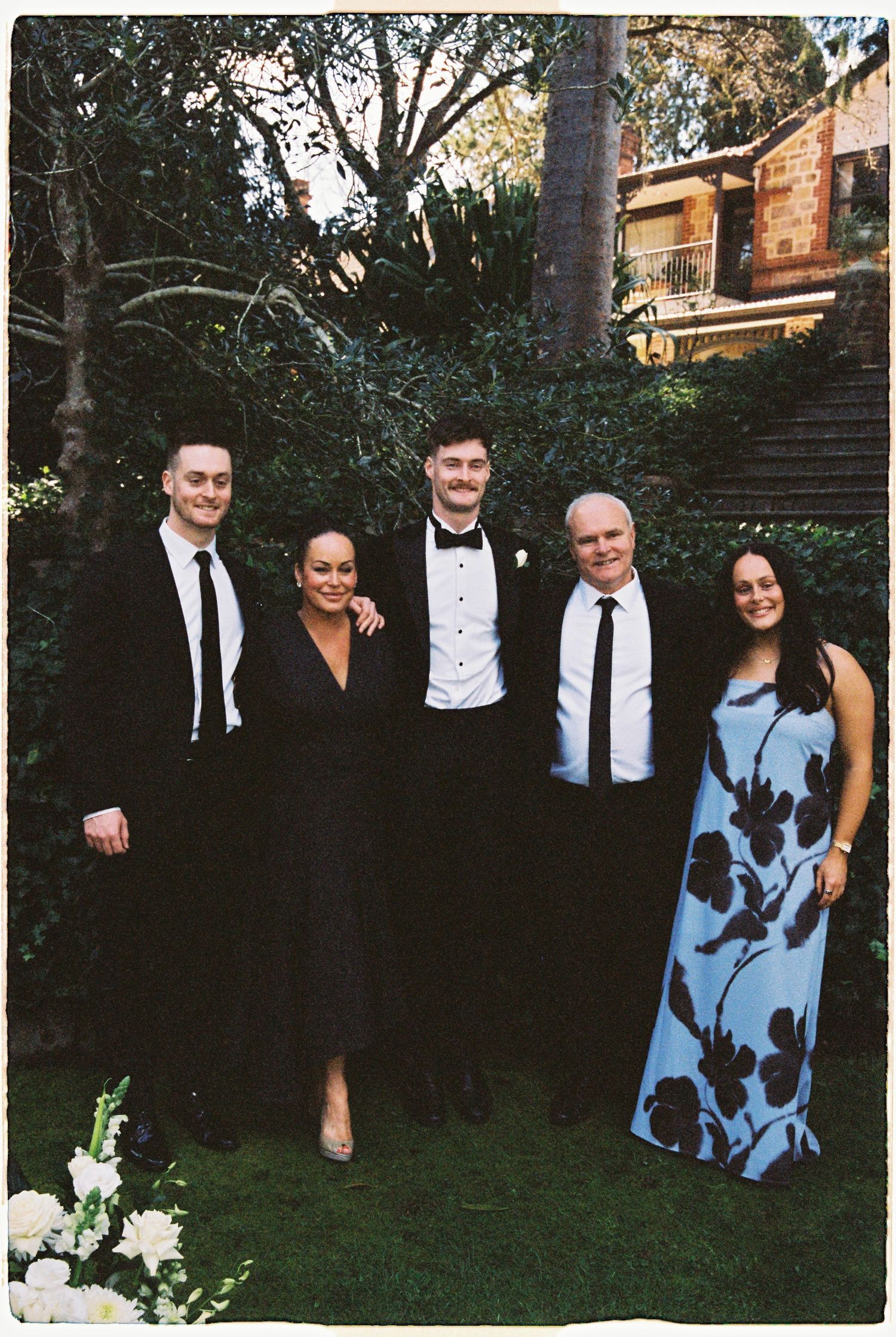 Five elegantly dressed wedding guests pose together in a lush garden setting.