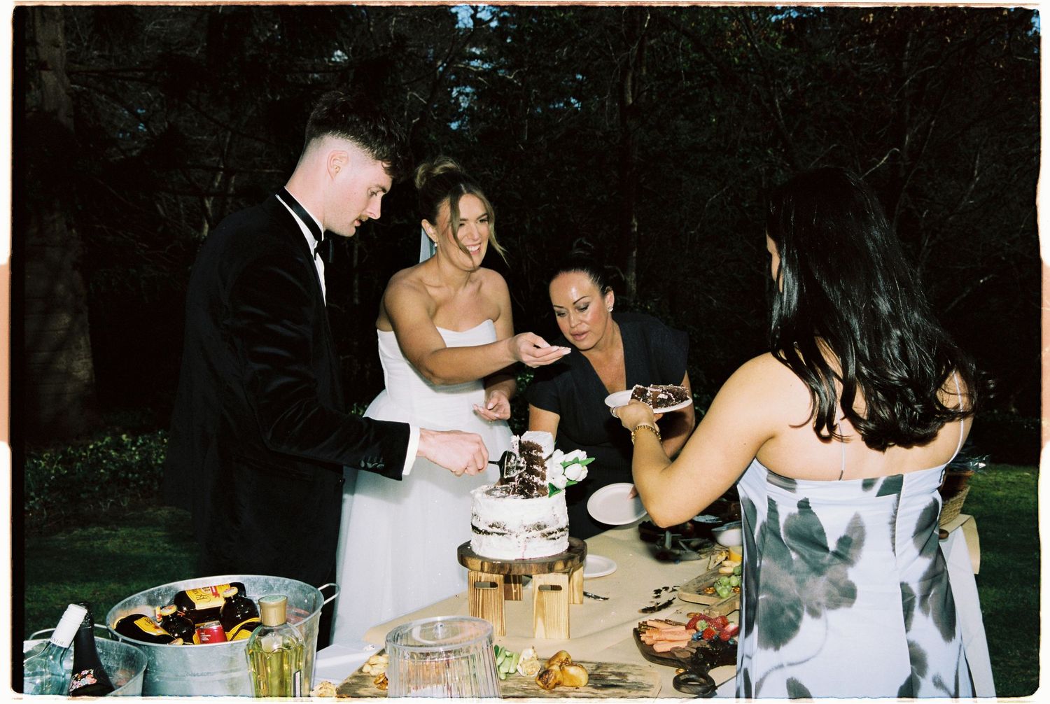 Bride and groom cut their wedding cake outdoors at night surrounded by guests and a dessert table.