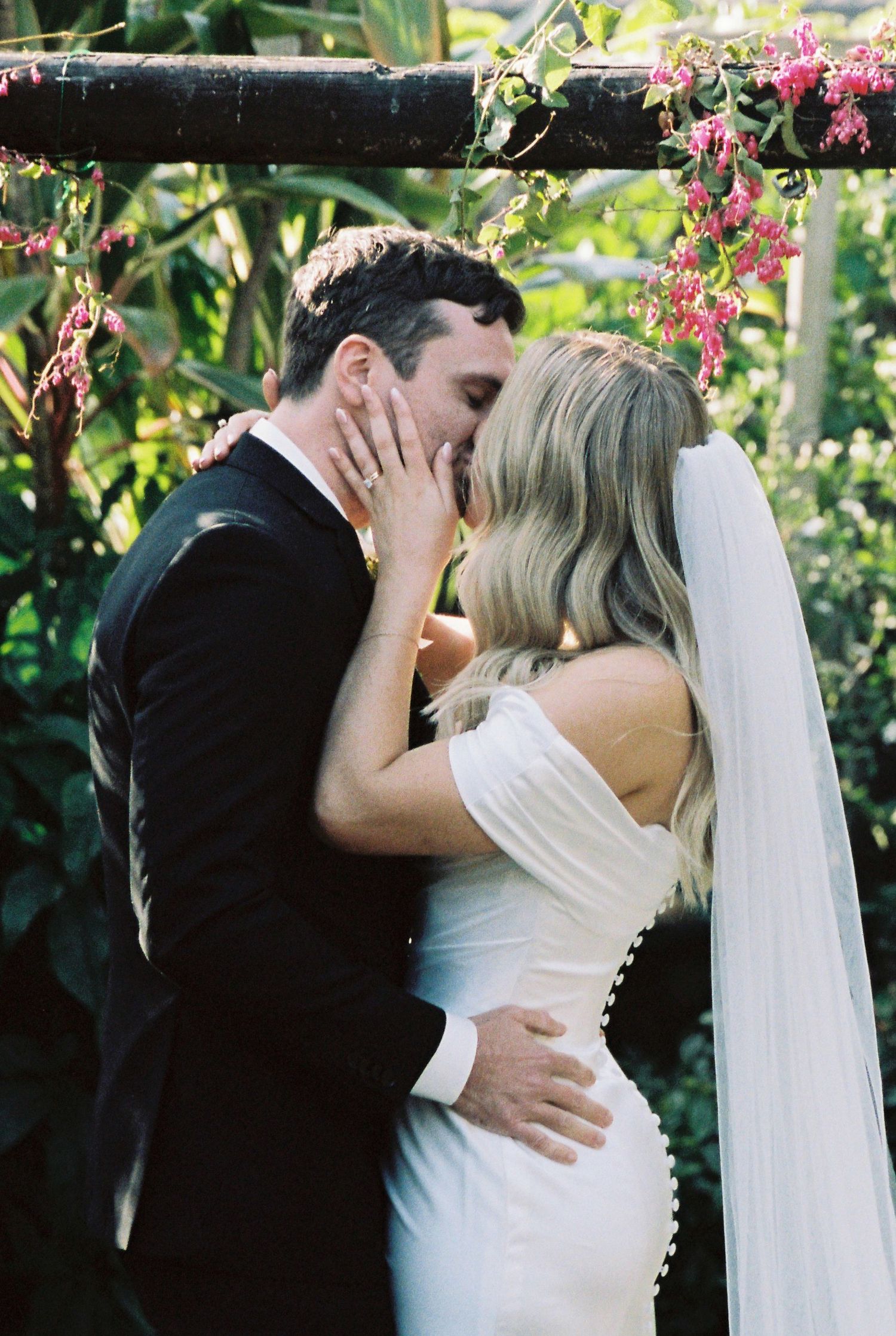 A bride and groom share their first kiss under a floral arch surrounded by lush greenery at an outdoor wedding ceremony.