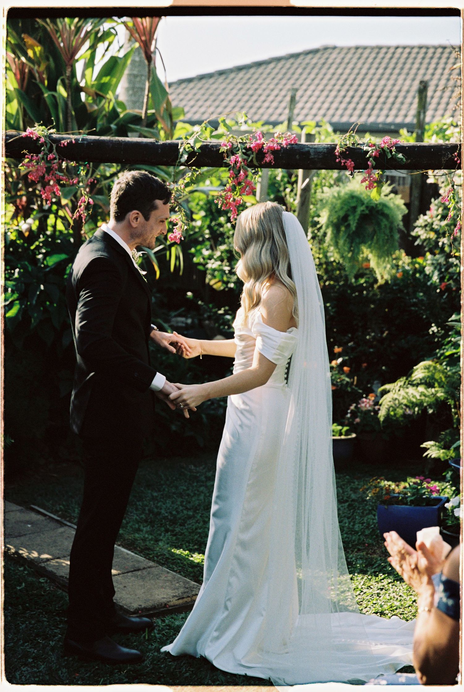 Bride and groom exchange vows in a lush garden ceremony, surrounded by tropical greenery and pink flowers.