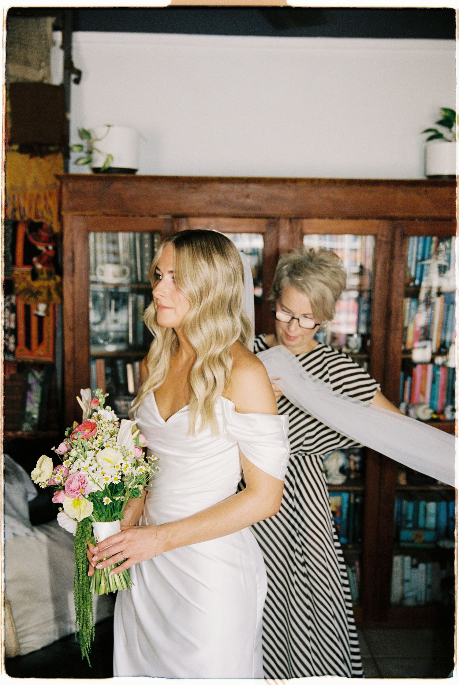 Older woman helping bride with off-shoulder white gown while bride holds colorful floral bouquet.