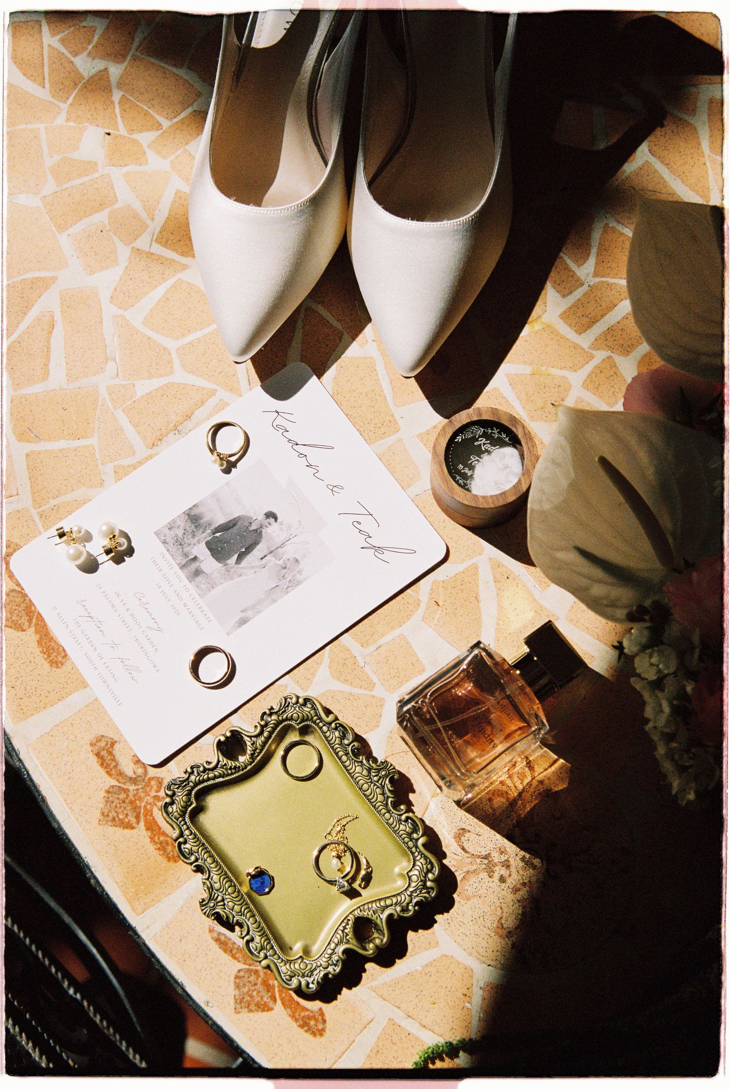 Overhead flat lay of bridal accessories including white heels, rings, perfume, and wedding invitation.