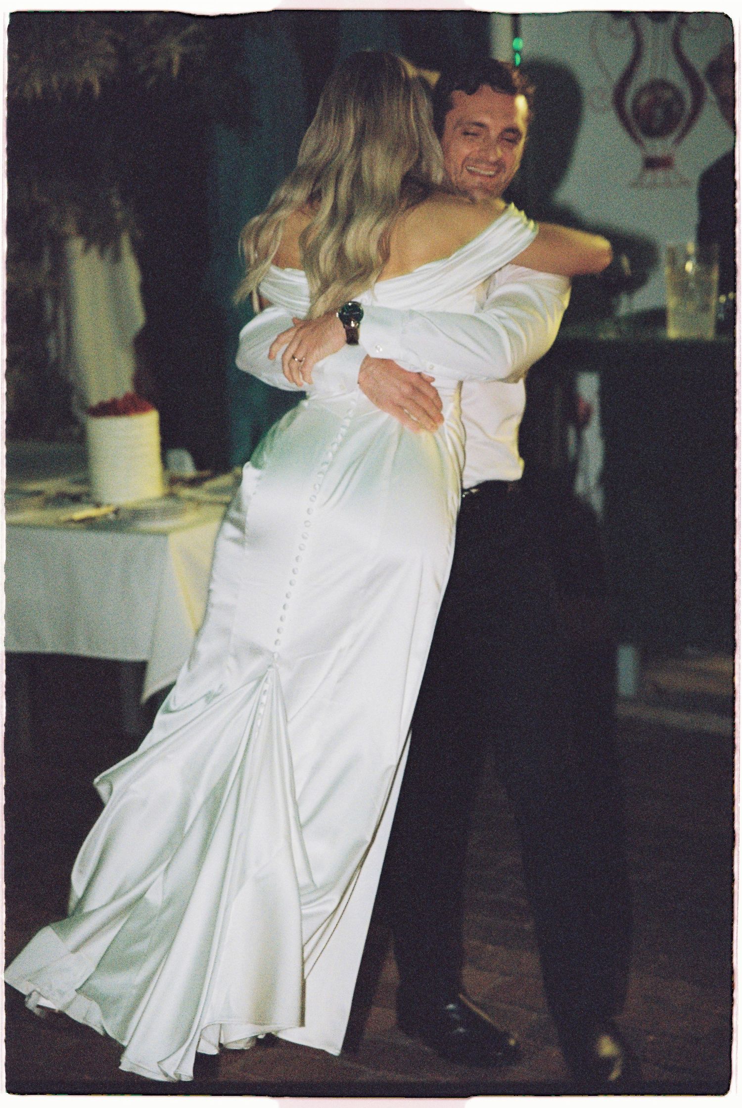 Bride and groom sharing first dance, bride in elegant white satin off-shoulder wedding gown with train.