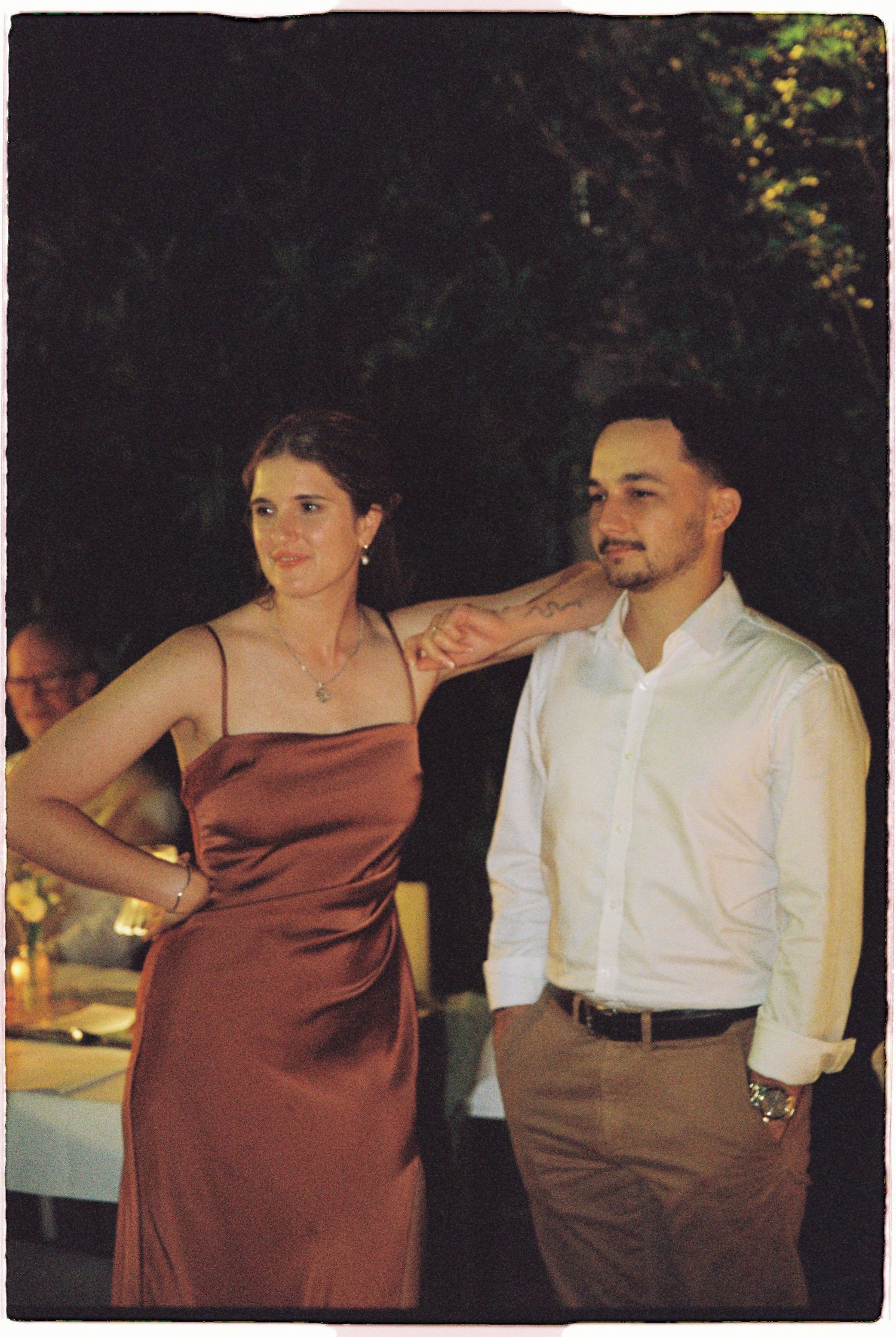 Elegant wedding guests, woman in rust satin slip dress and man in white shirt, posing at reception.