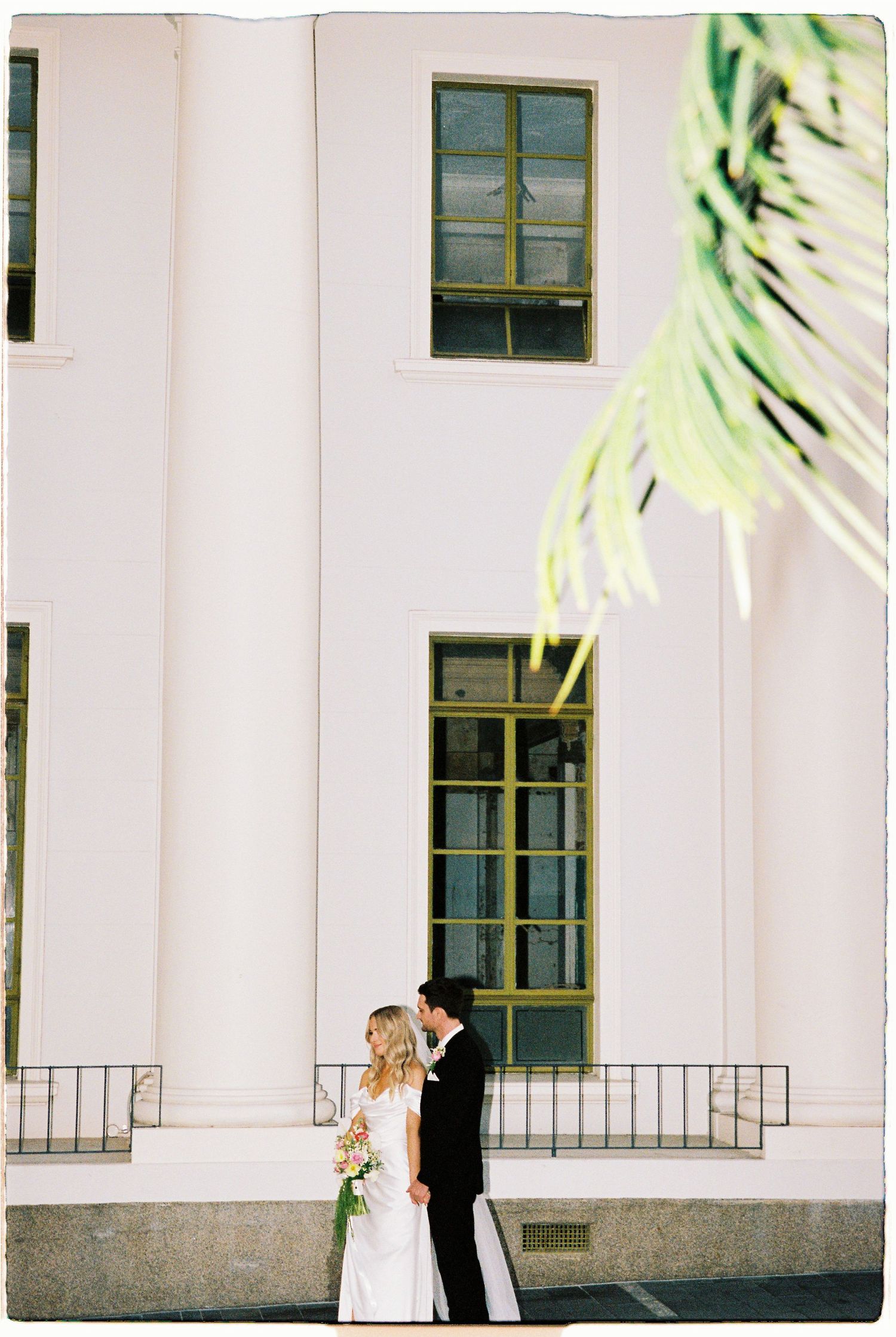 Bride and groom stand close together before a grand white classical building framed by tropical palm fronds.