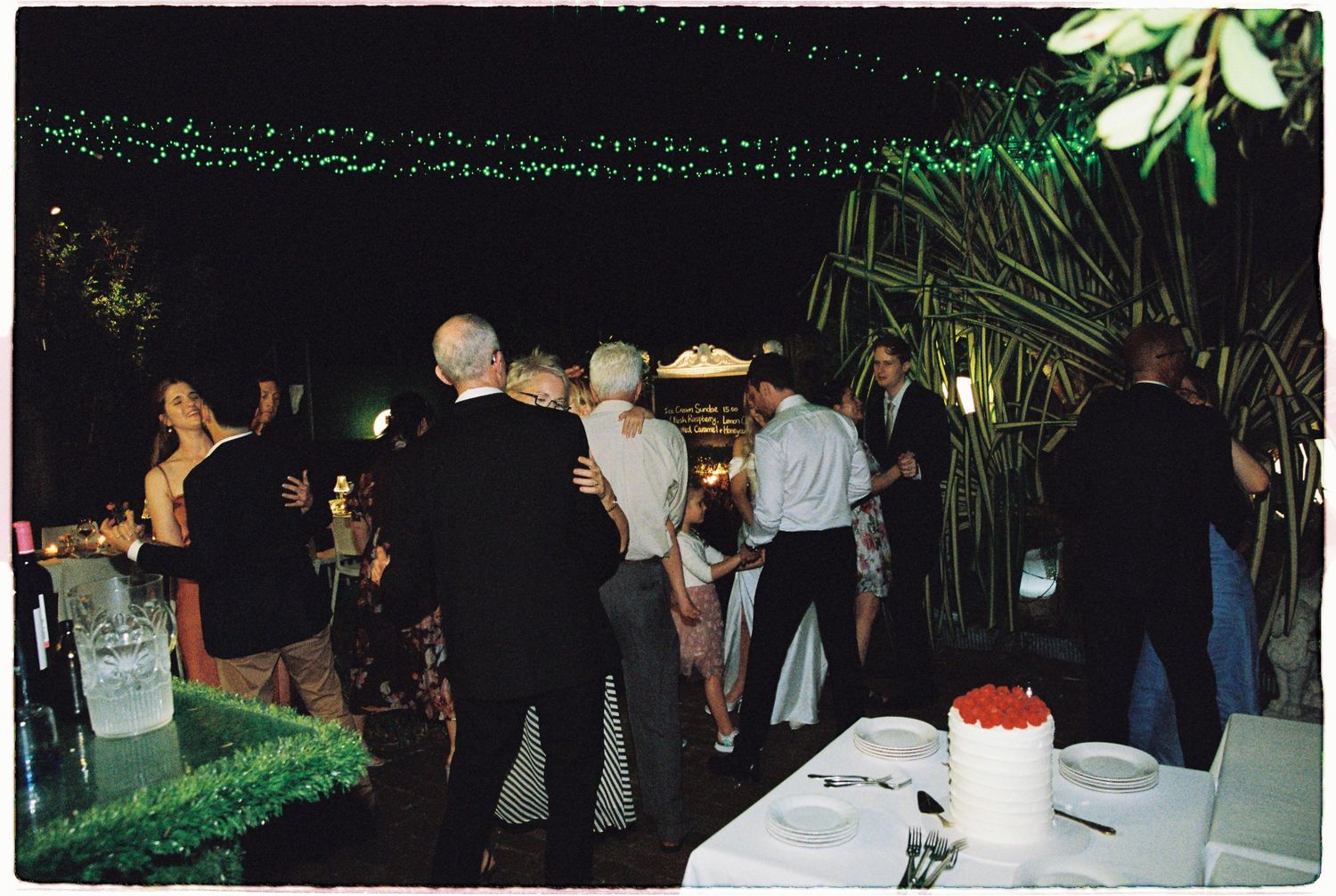 Guests mingle at an outdoor evening party with string lights, tropical plants, and a decorated table in the background.