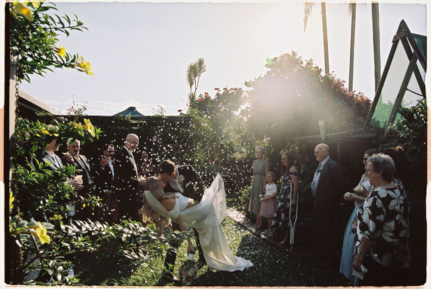 Bride and groom kissed with confetti shower at sunlit outdoor garden wedding ceremony with guests.