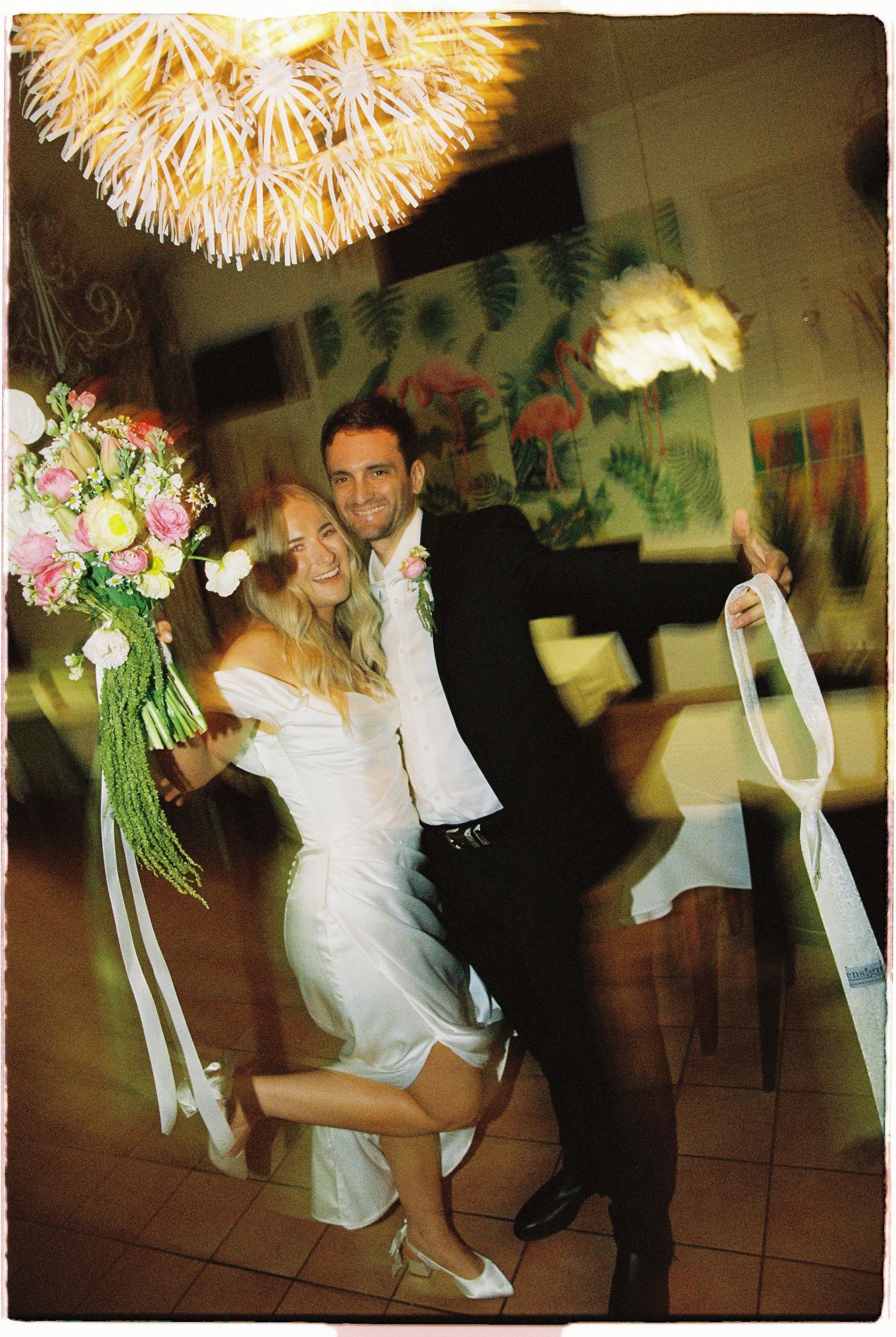 Joyful newlyweds dancing at wedding reception, bride holding pink floral bouquet with ribbons.