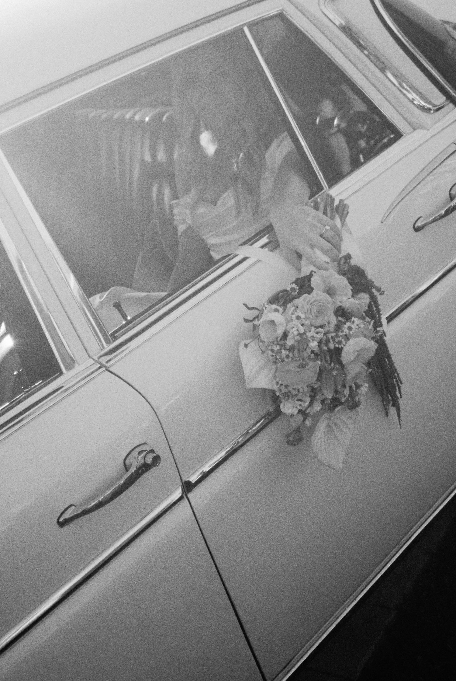 Bride holding floral bouquet beside a vintage classic car, black and white wedding photo.