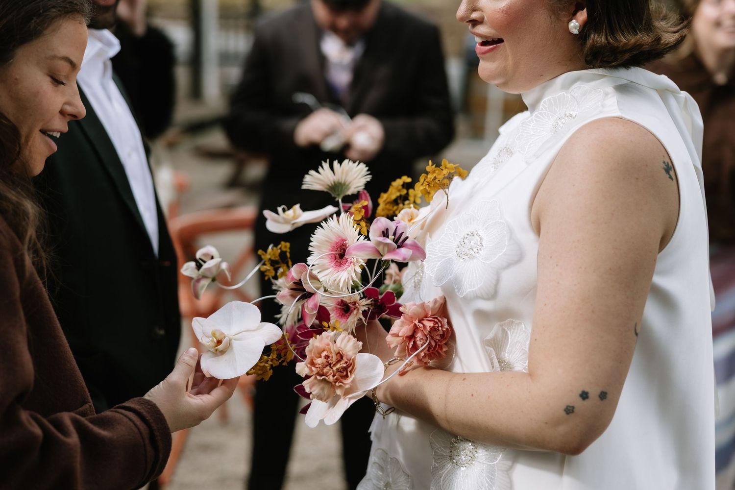 Bride in white dress holds colorful wedding bouquet with pink, white, and yellow flowers while guest admires blooms.
