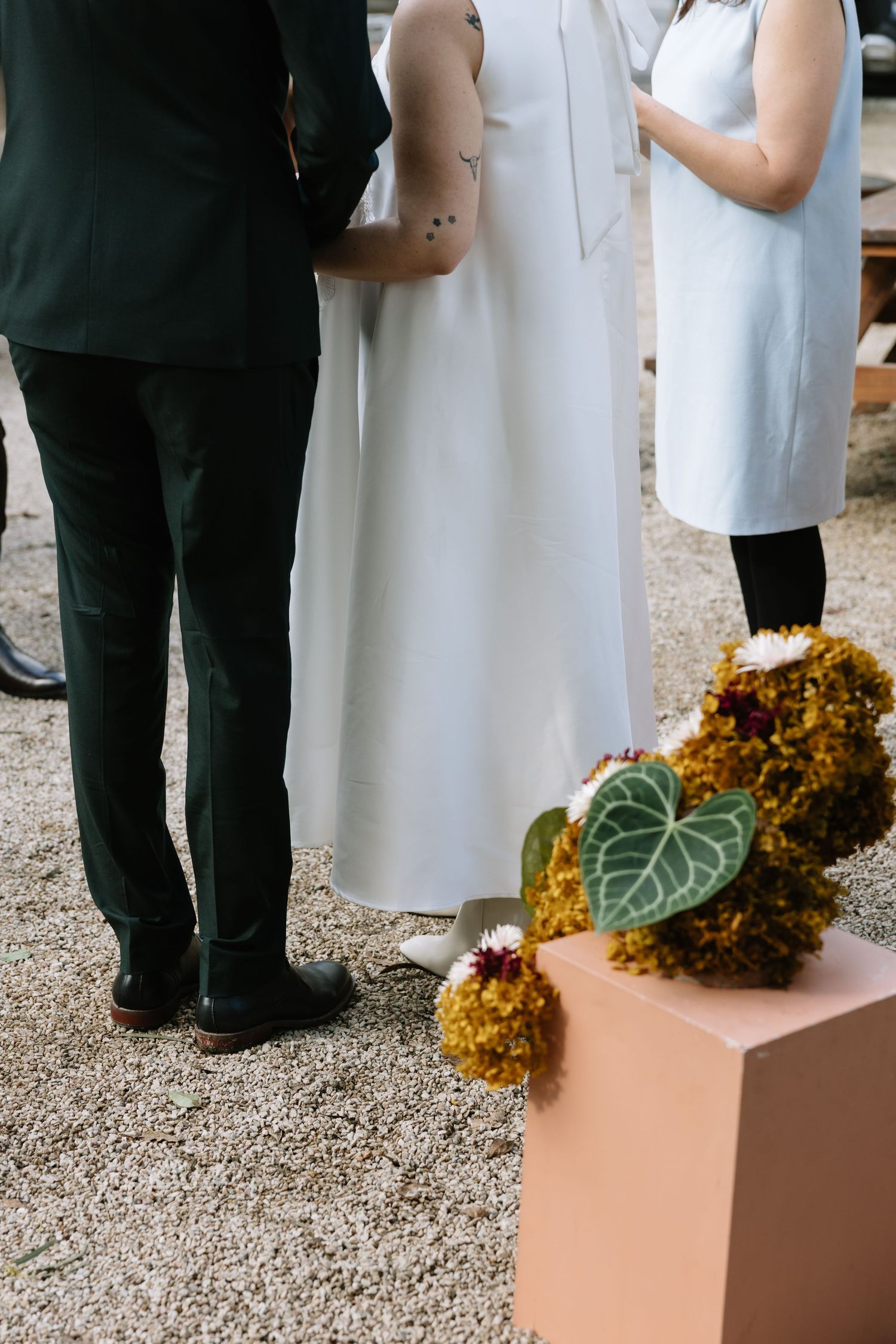 Wedding ceremony with couple in white and dark green, officiant, and floral arrangement on a pink pedestal.