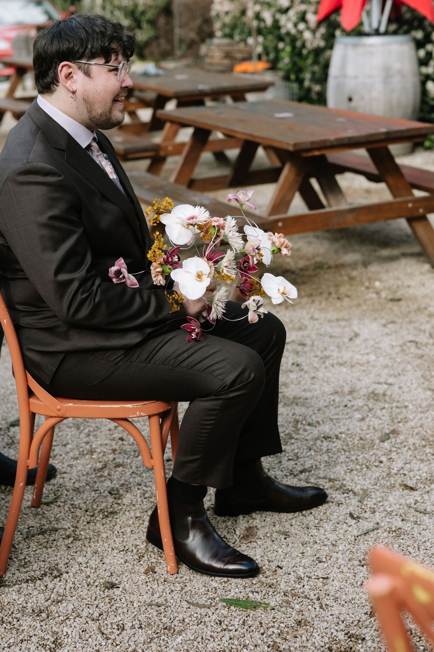 A groom in a dark suit sits on an orange chair holding a floral bouquet at an outdoor wedding venue.