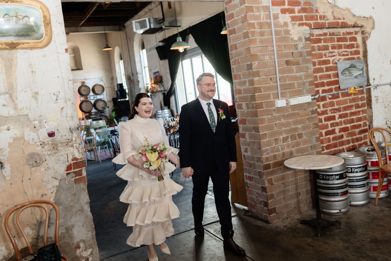 Bride and groom walk through rustic brick wedding venue with exposed walls and beer kegs.
