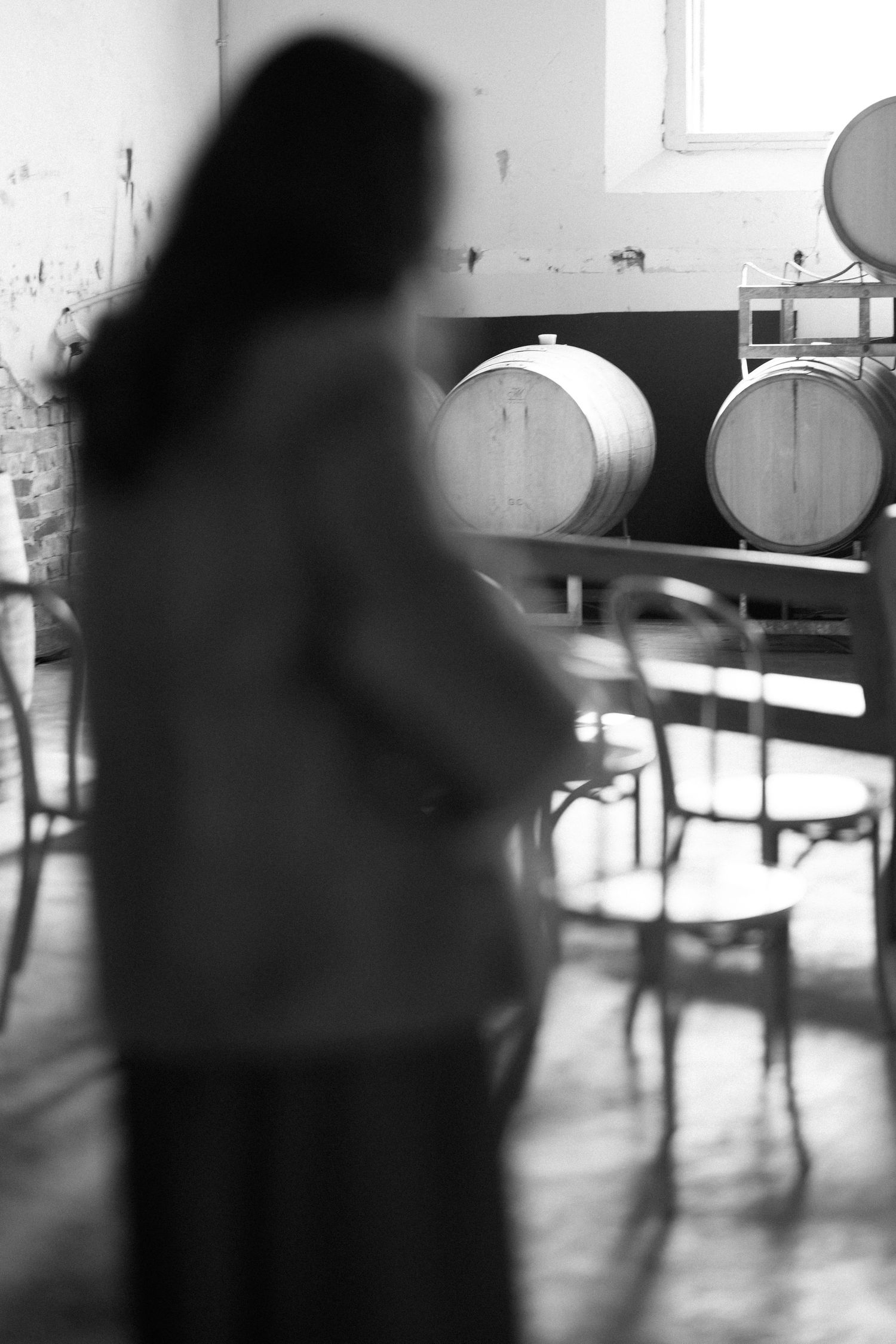 Black and white photo of a woman standing near wooden wine barrels in a rustic winery tasting room.