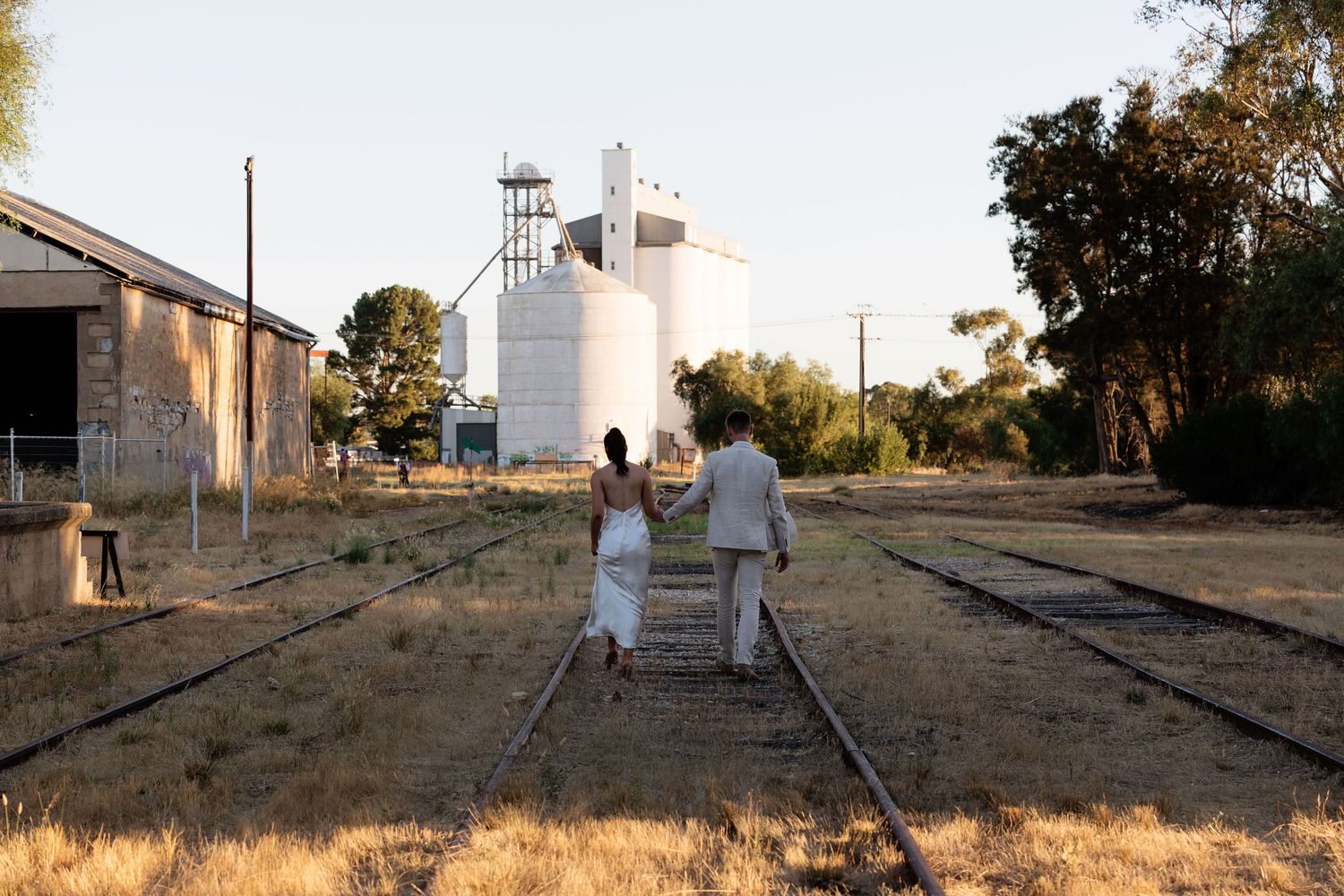 Couple walks hand-in-hand along railway tracks toward grain silos in a rural Australian town at golden hour.