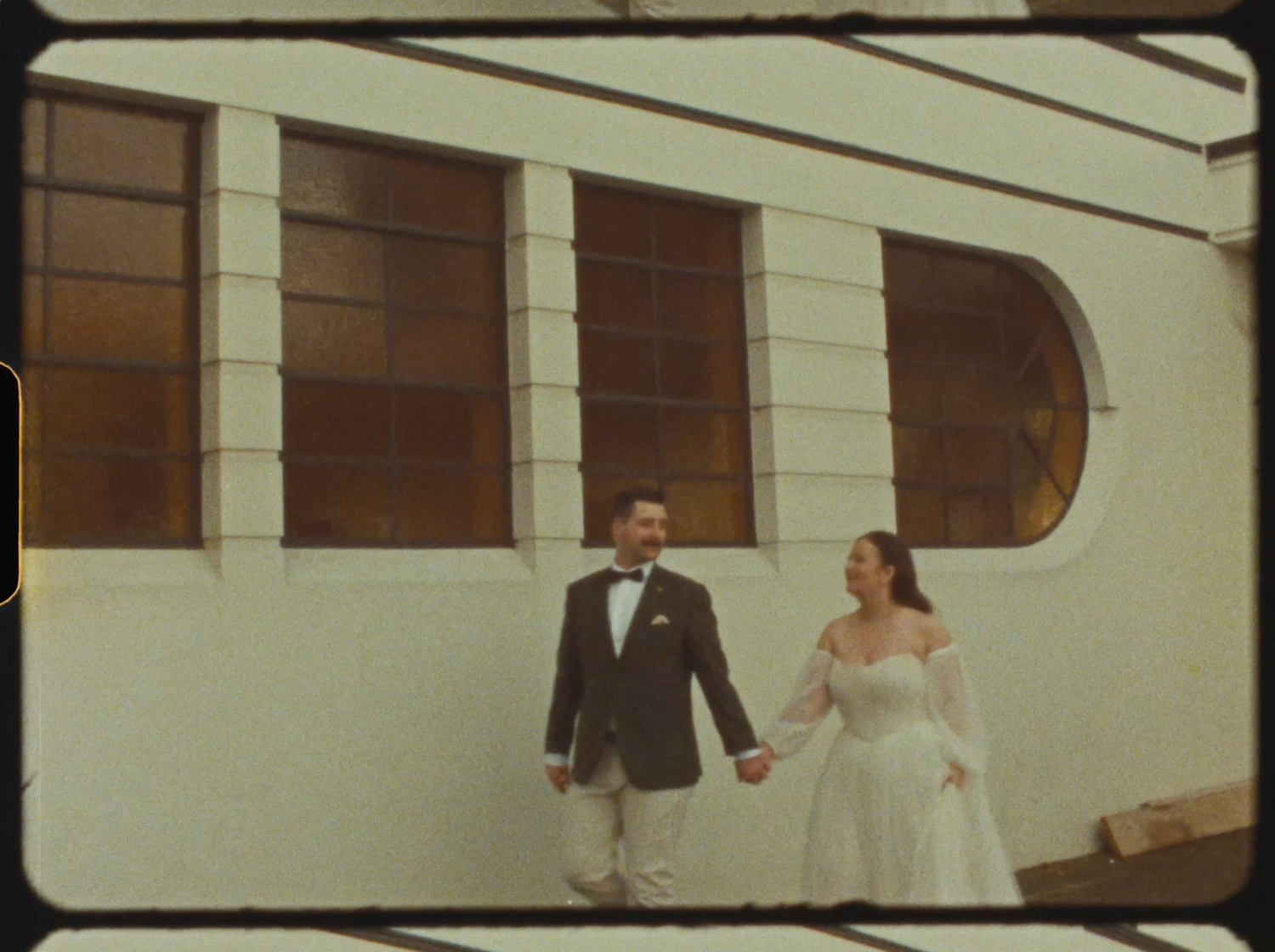 A bride and groom hold hands walking outside a white building with arched windows on their wedding day.