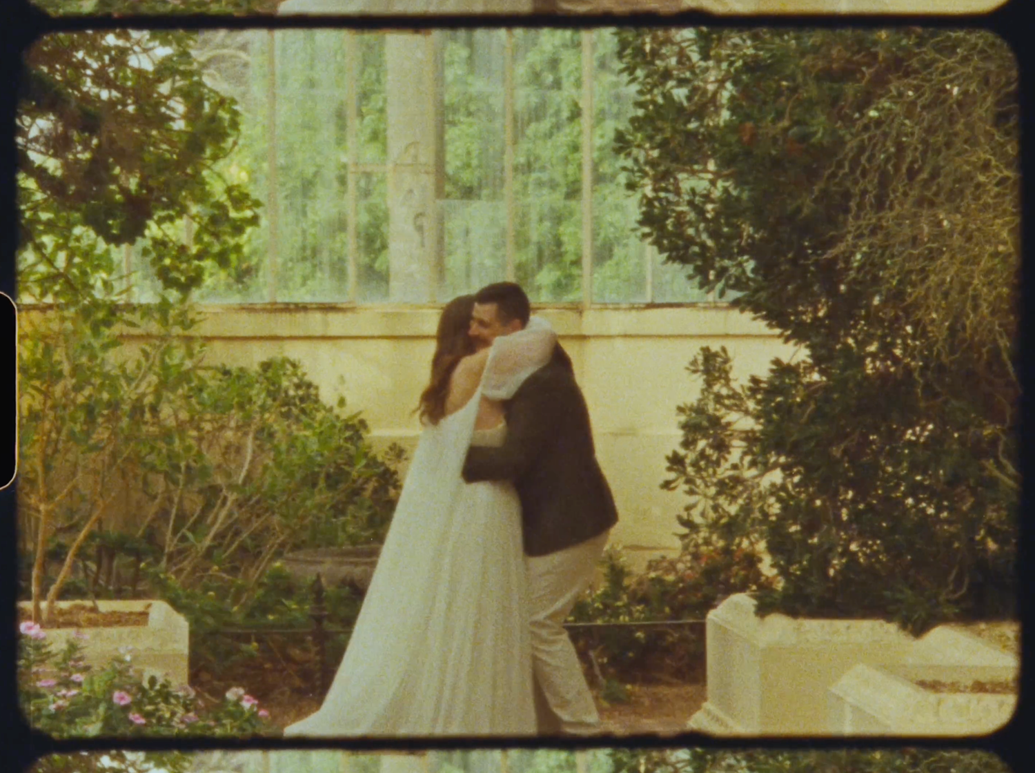 A bride and groom embrace tenderly in a lush garden greenhouse wedding setting.