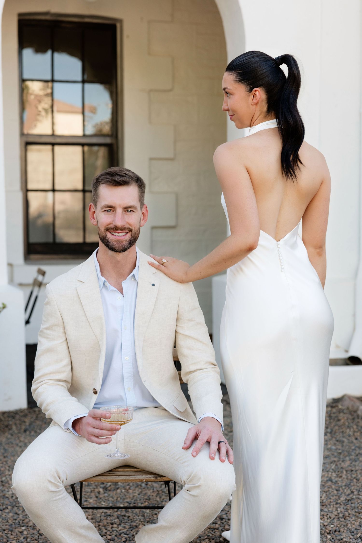 Smiling groom seated outdoors admires bride in backless white satin halter wedding dress.