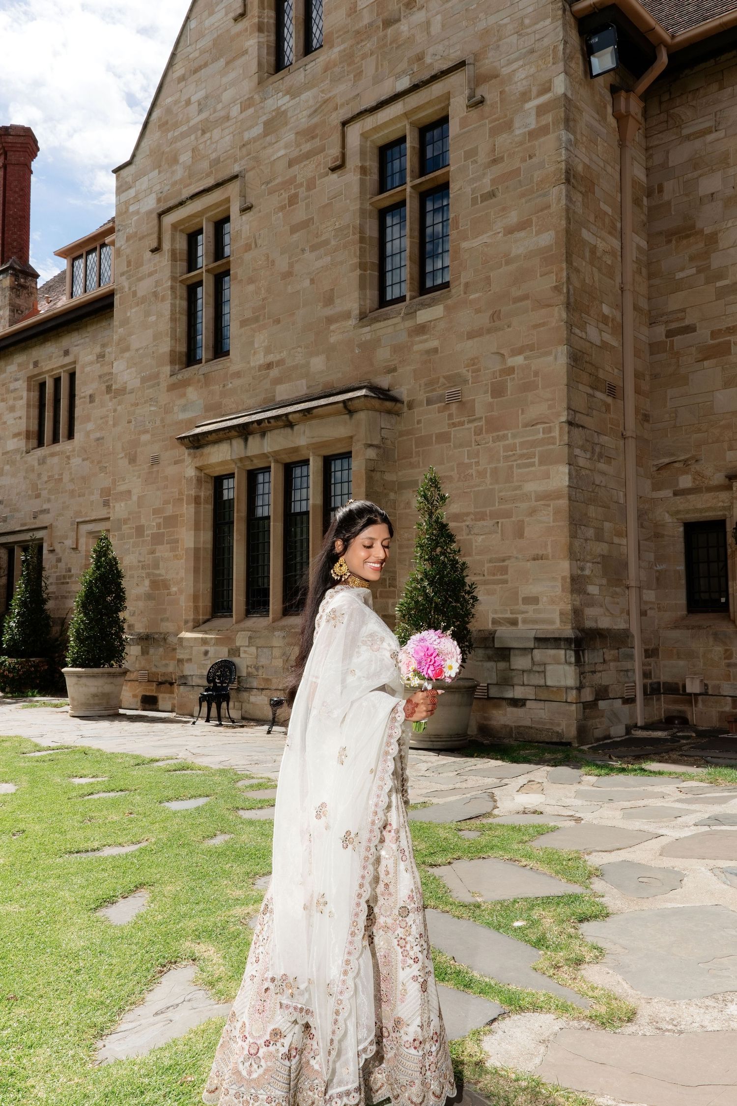 South Asian bride in ornate white lehenga with dupatta, holding pink bouquet beside elegant stone manor.