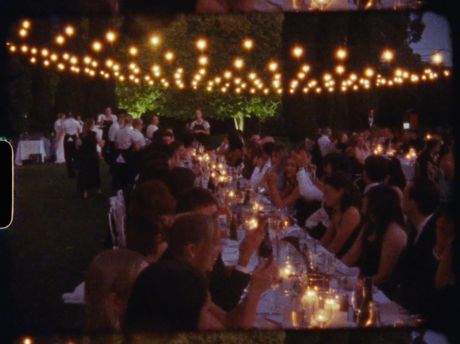 Guests seated at long candlelit tables under string lights at nighttime outdoor wedding reception.