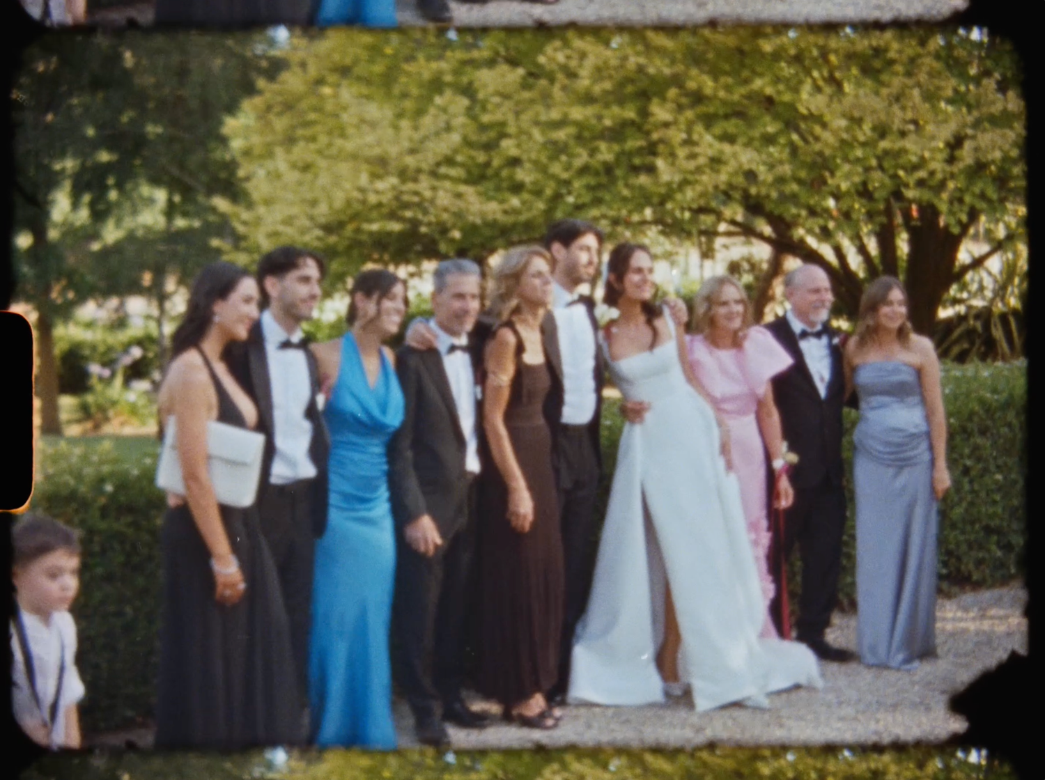 Large wedding party group photo in formal attire posing in a sunny garden with lush green hedges.