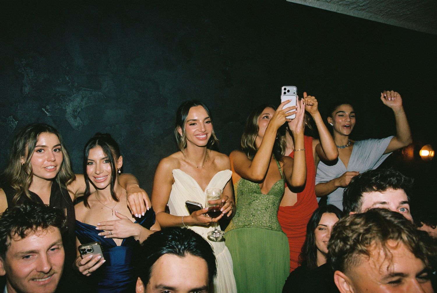 Smiling bride holding drink surrounded by celebrating guests at wedding party.