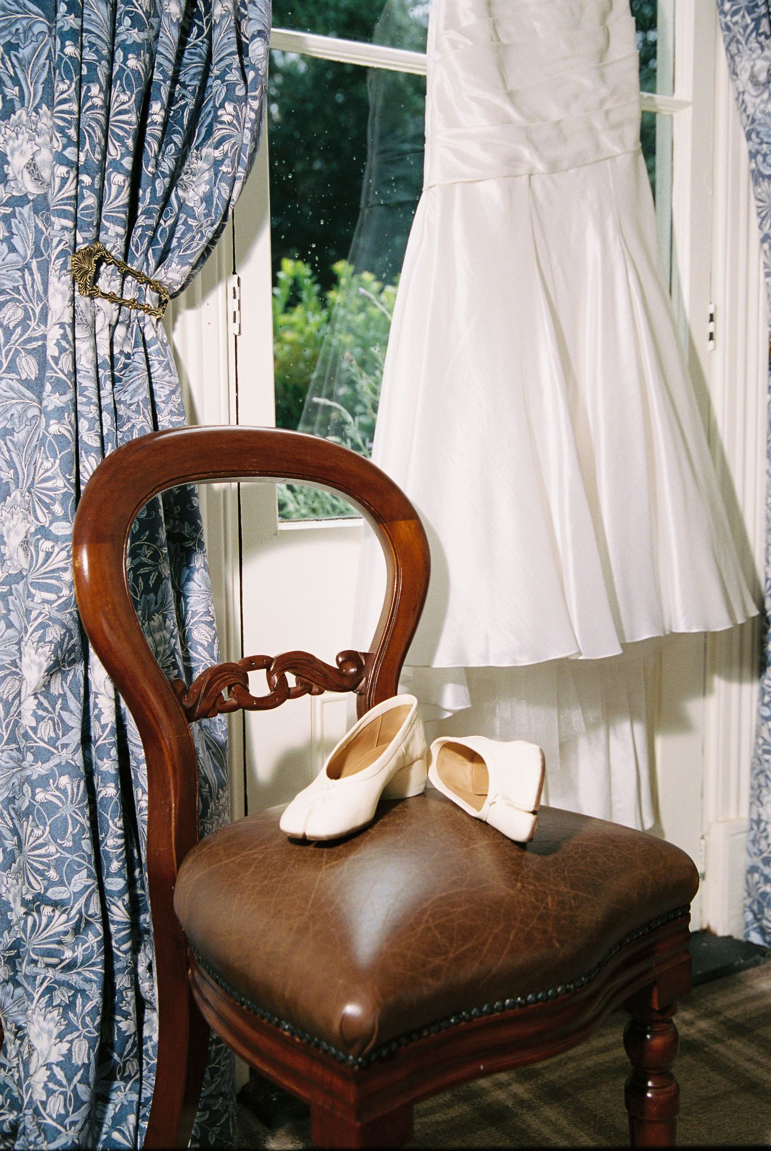 White wedding gown hanging in window with white ballet flat bridal shoes on antique wooden chair.