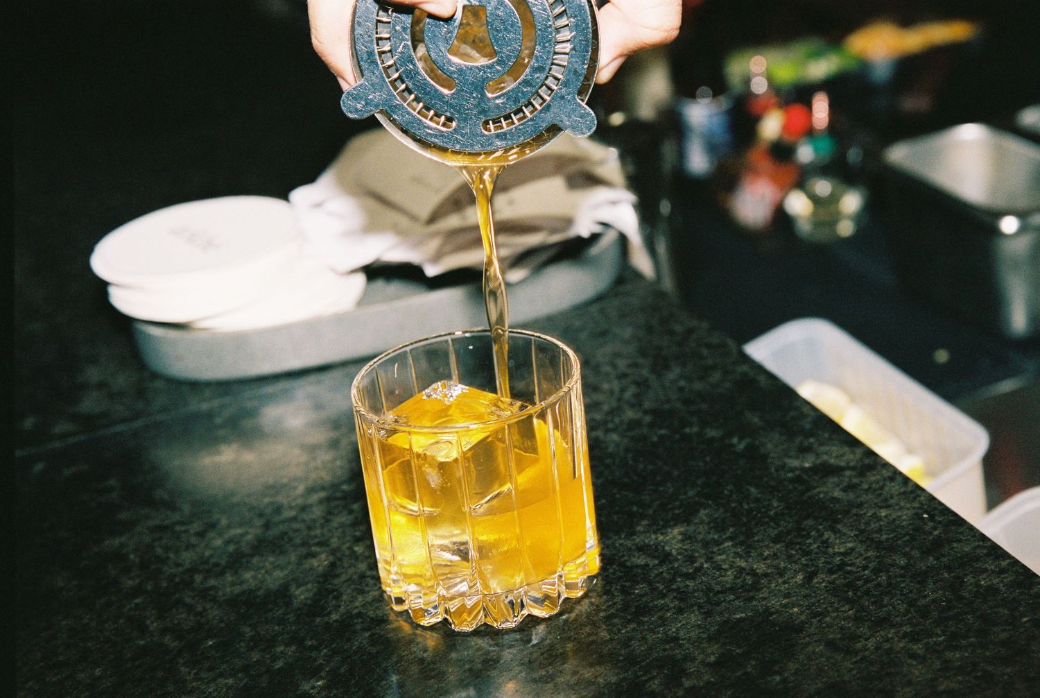 Bartender straining amber cocktail over large ice cube into rocks glass on dark marble bar surface.