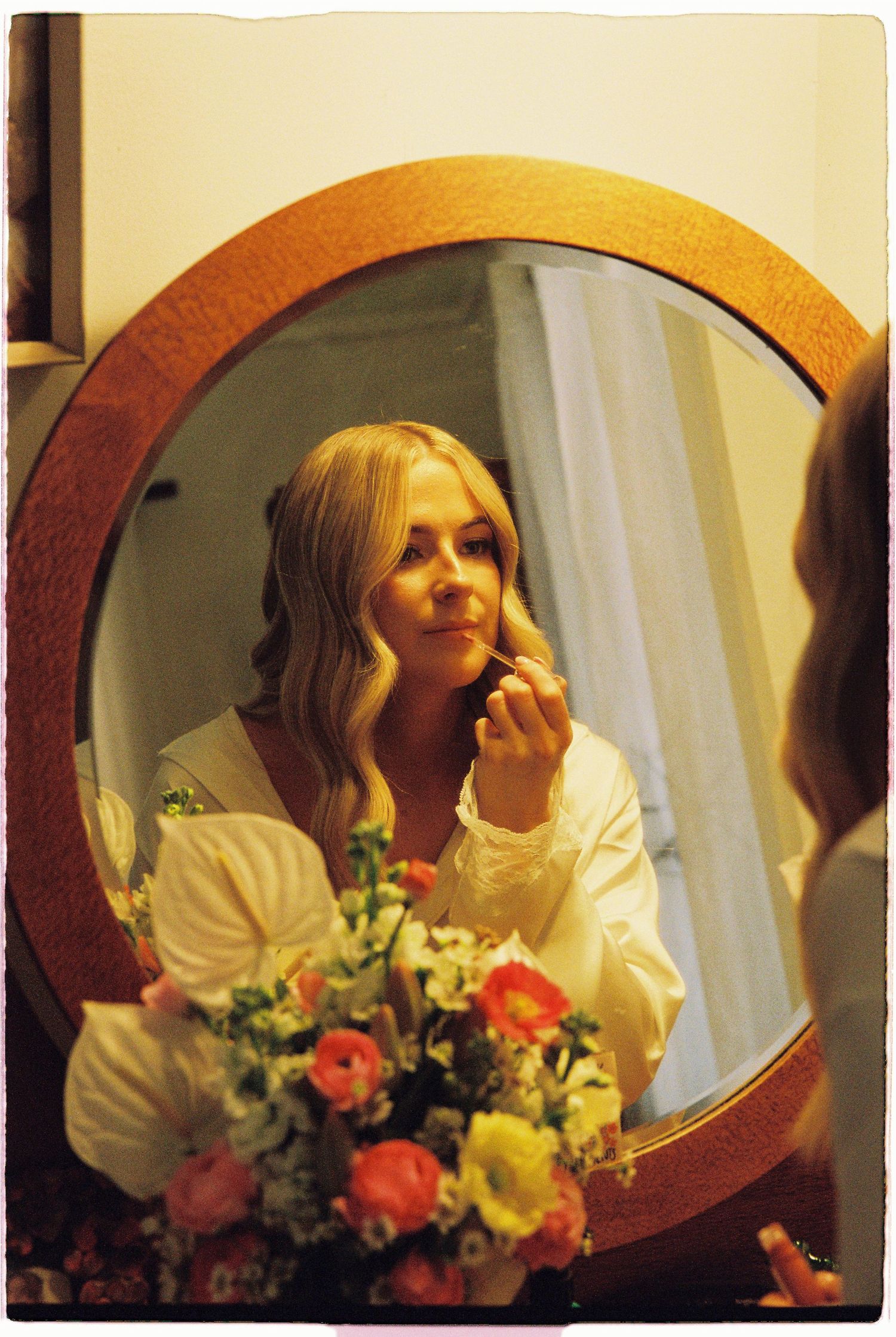 Blonde bride applies makeup at round wooden mirror surrounded by colorful wedding flowers.