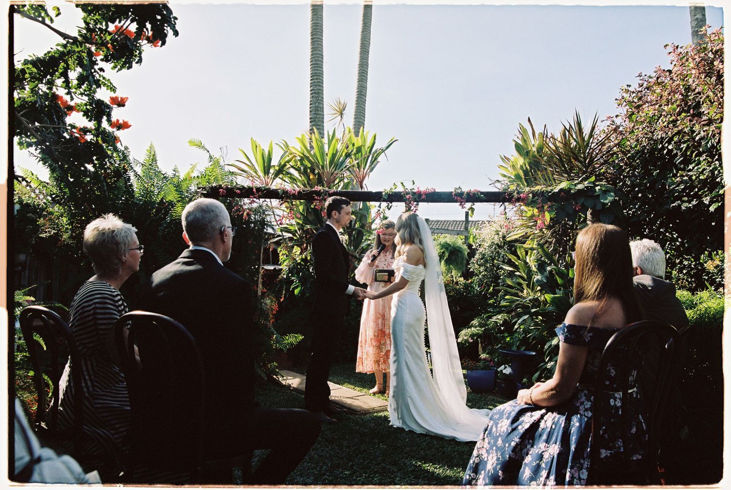 Outdoor garden wedding ceremony with bride and groom at floral arch surrounded by guests.
