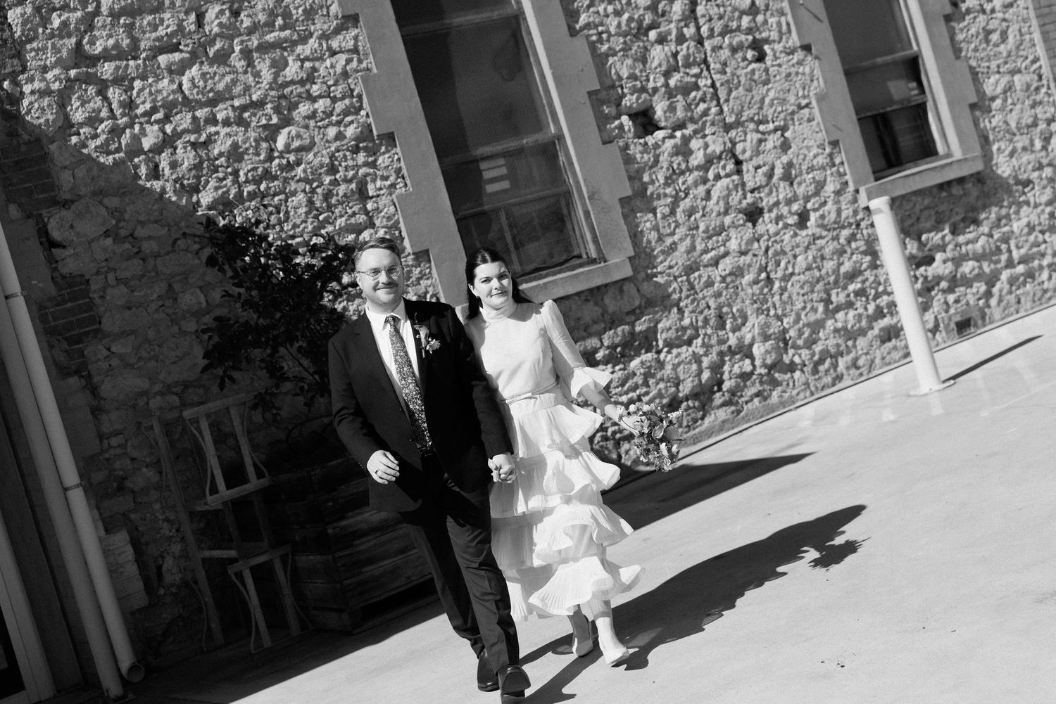Black and white photo of bride in ruffled dress and groom walking beside historic stone building.