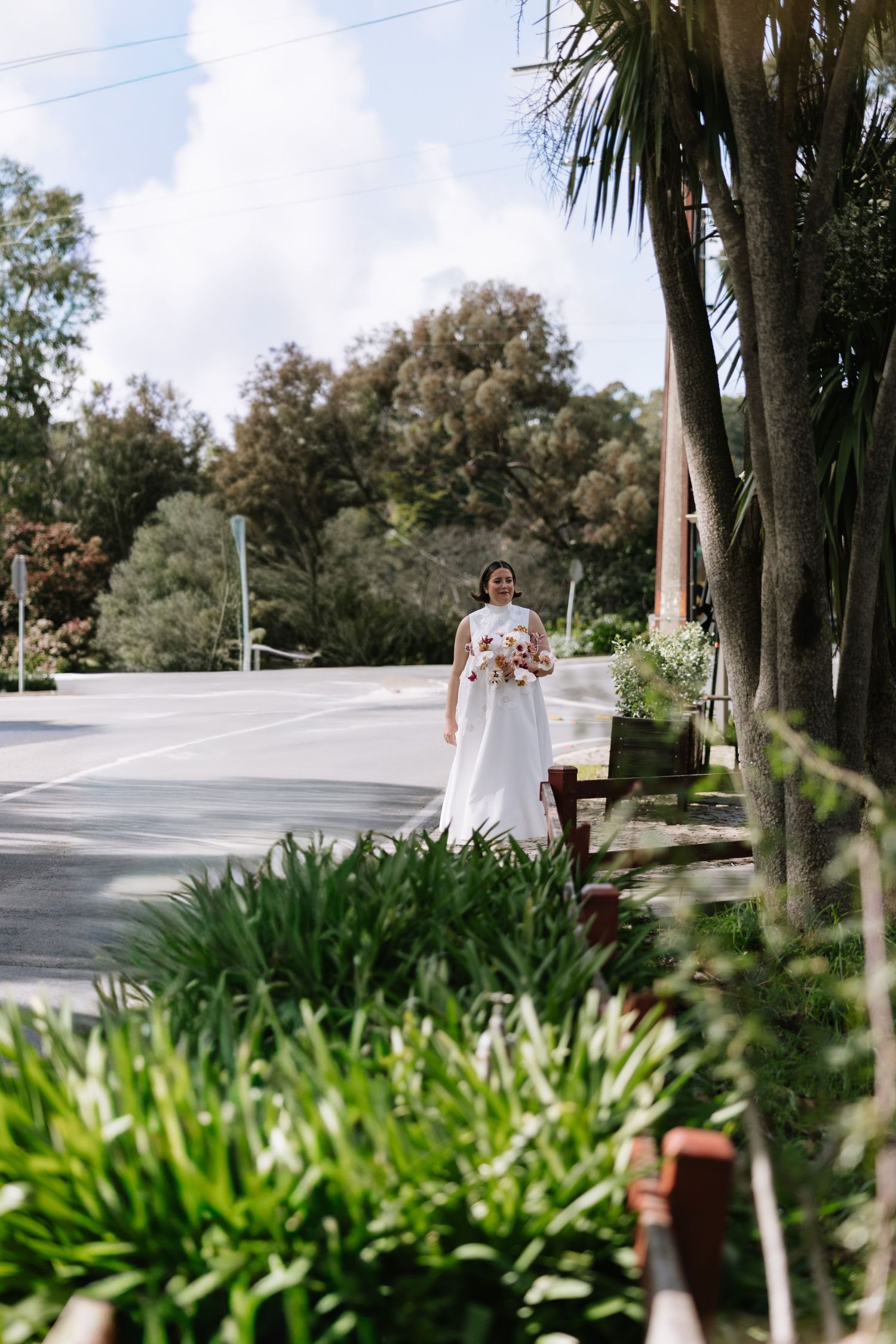 Bride in white gown holding bouquet standing on tree-lined street on sunny wedding day.