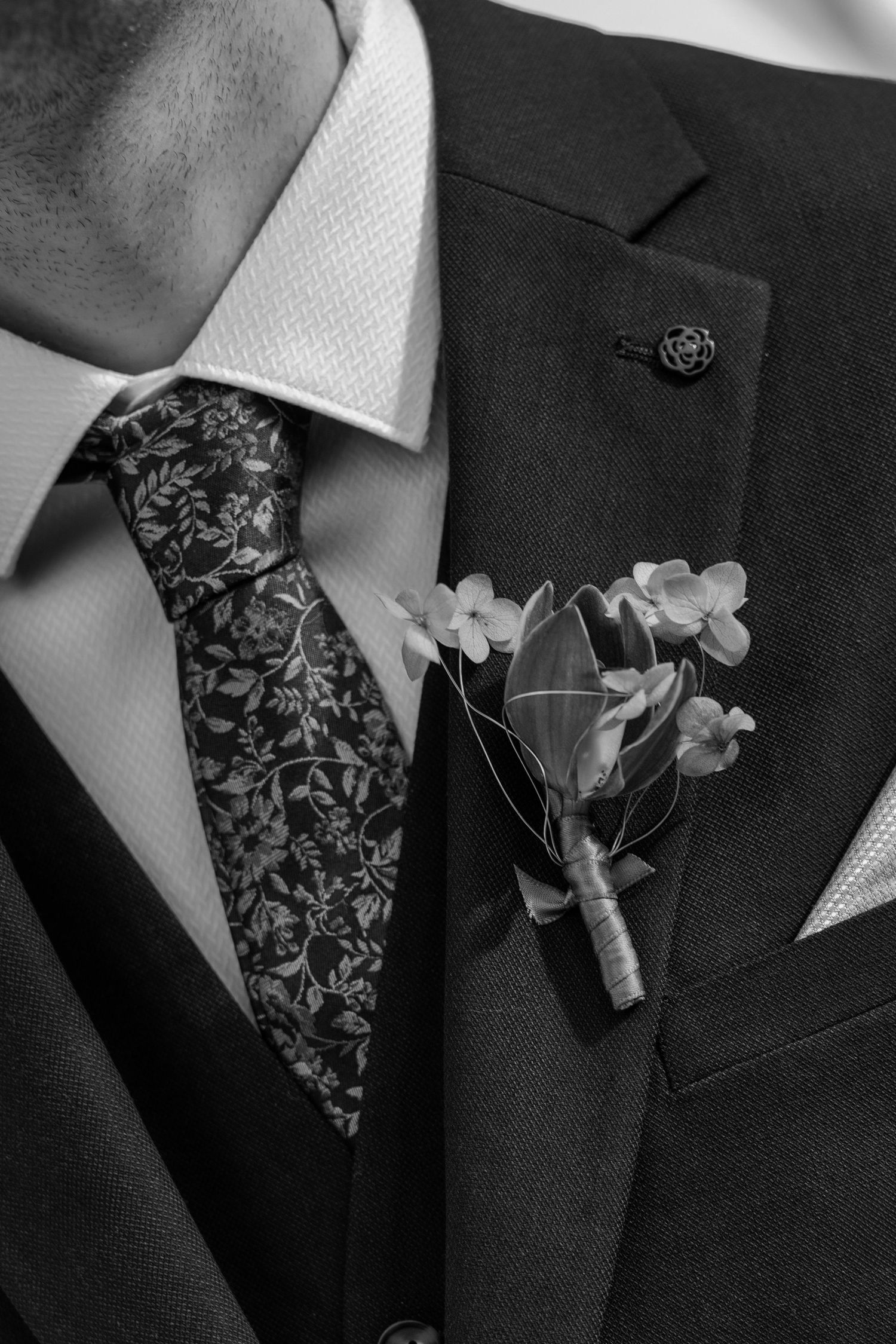 Black and white close-up of a groom wearing a floral tie, suit, and boutonniere with small white flowers.