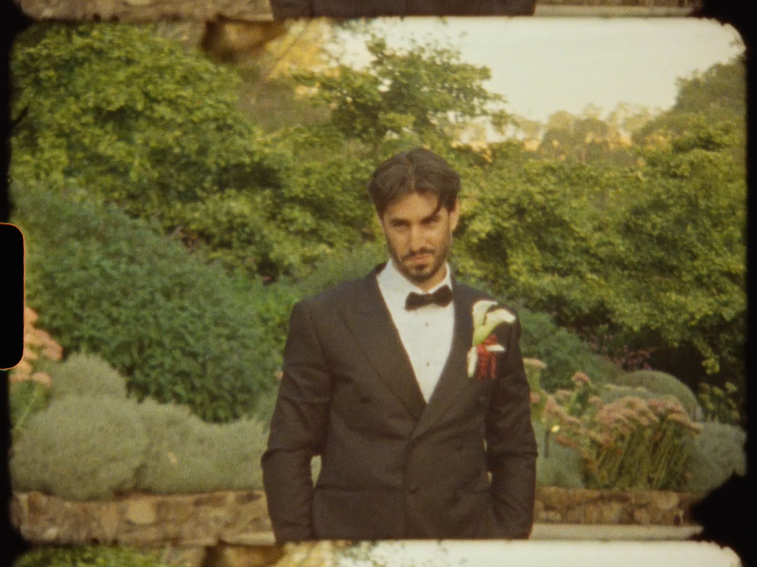 Groom in black tuxedo with bow tie and boutonniere stands in garden.