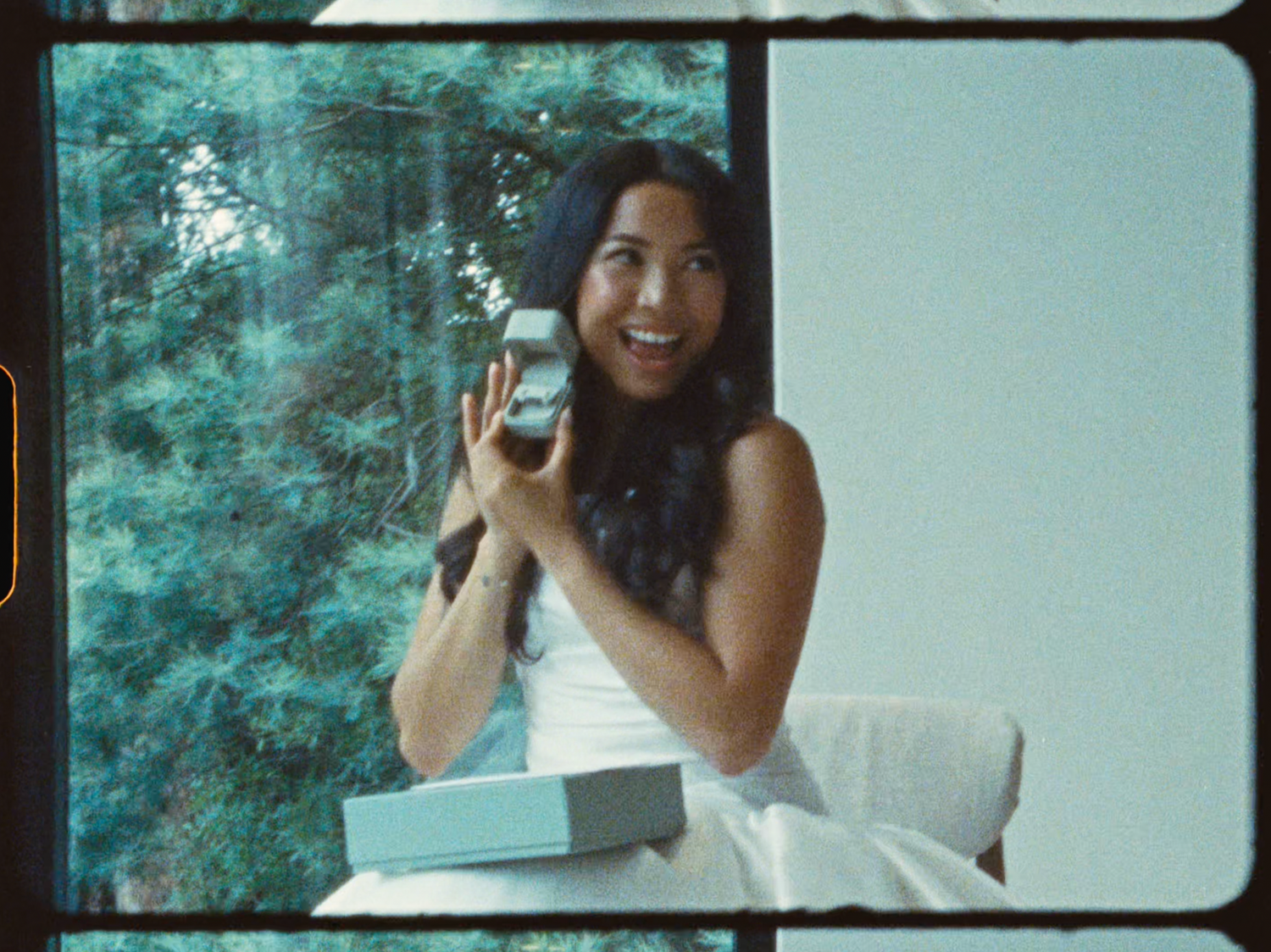 Smiling bride in white holds a camera while seated near large windows surrounded by greenery.