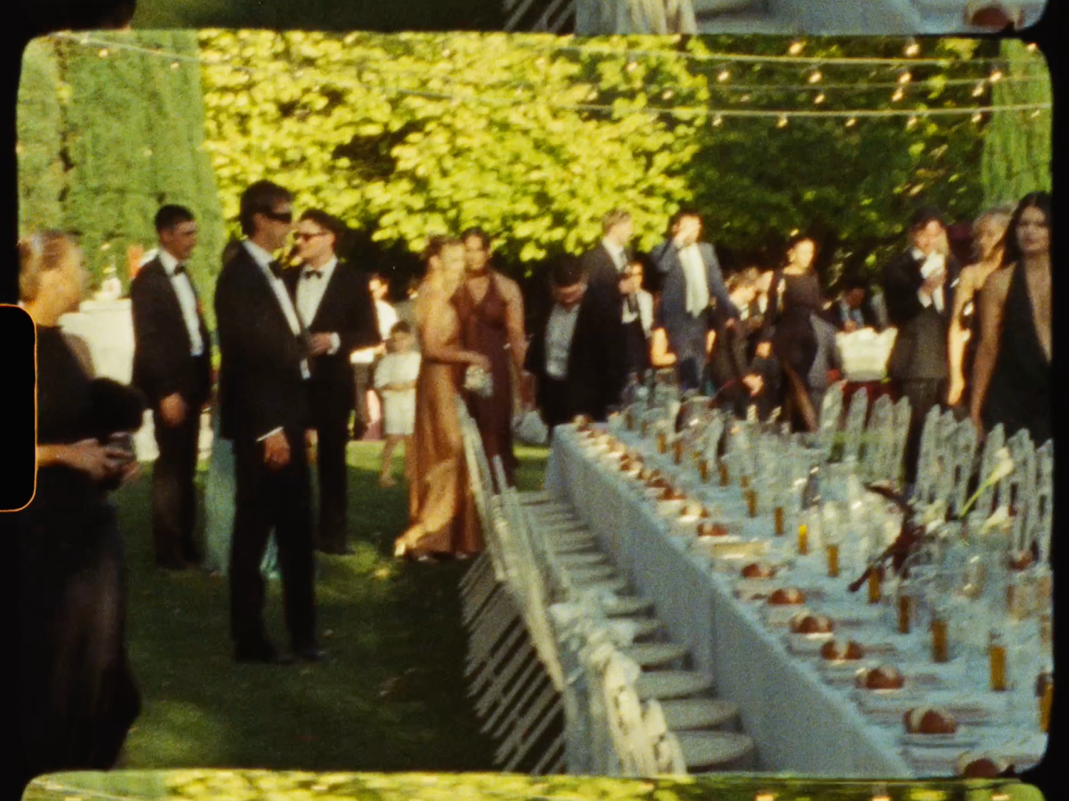 Wedding guests in formal attire mingling beside a long elegantly set outdoor reception table.