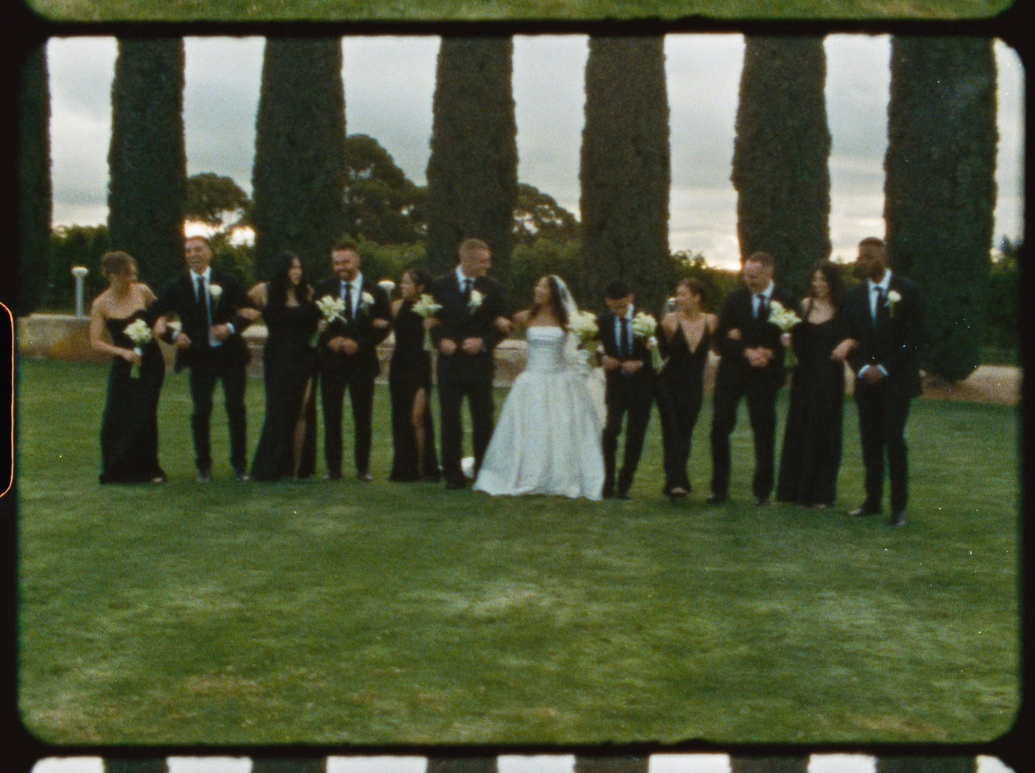 Elegant wedding party in black formal wear flanking bride and groom on lush green lawn at sunset.