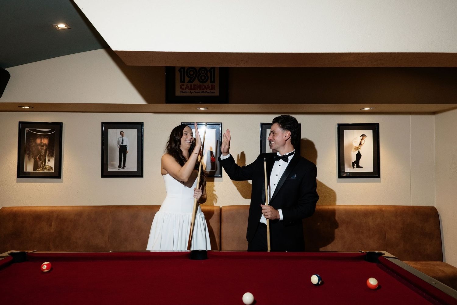 Bride and groom in formal wear laughing together while holding pool cues beside red billiard table.